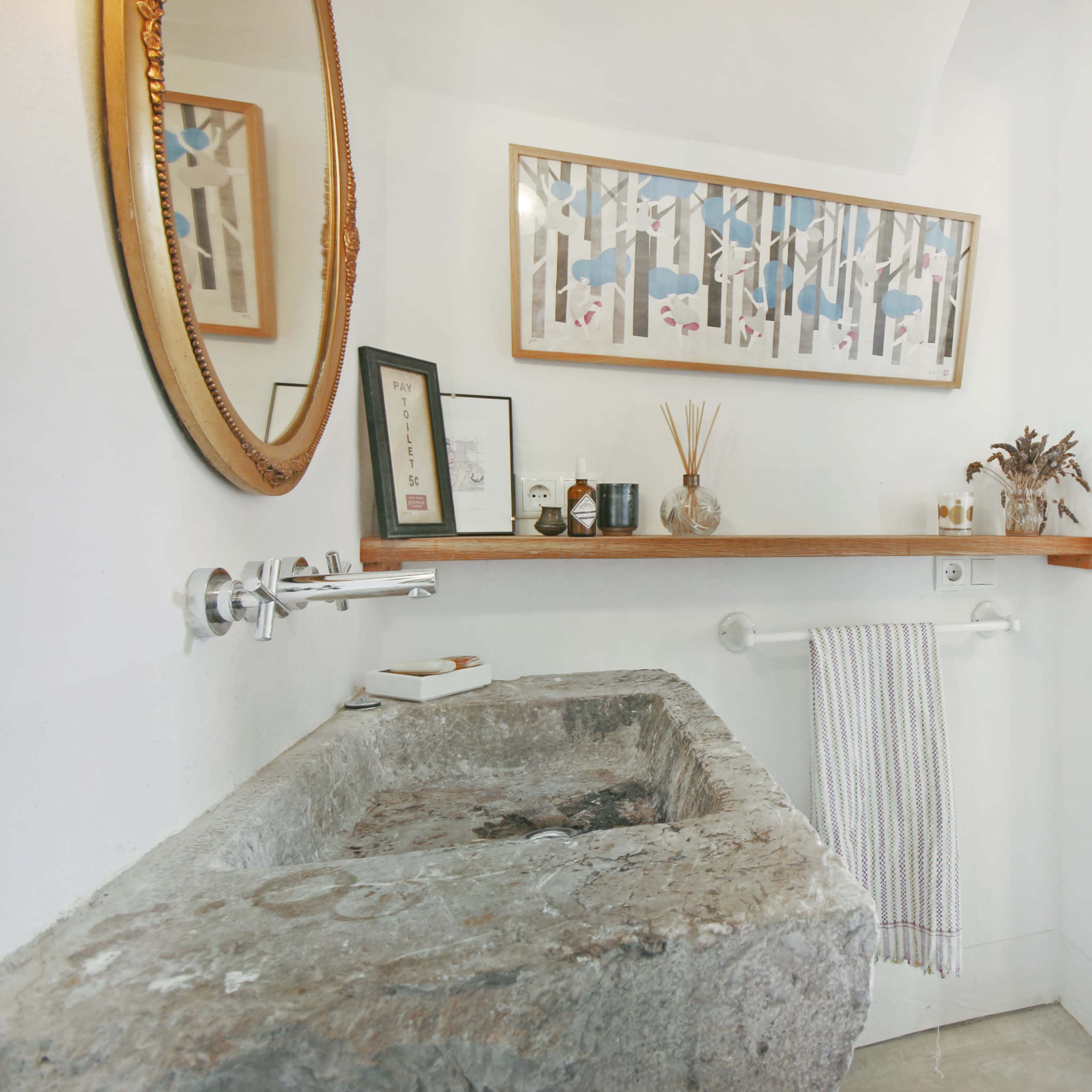 A minimalist bathroom featuring a carved stone sink, a wooden shelf with various items, and a doorway leading to a brightly lit room with a patterned bedspread.