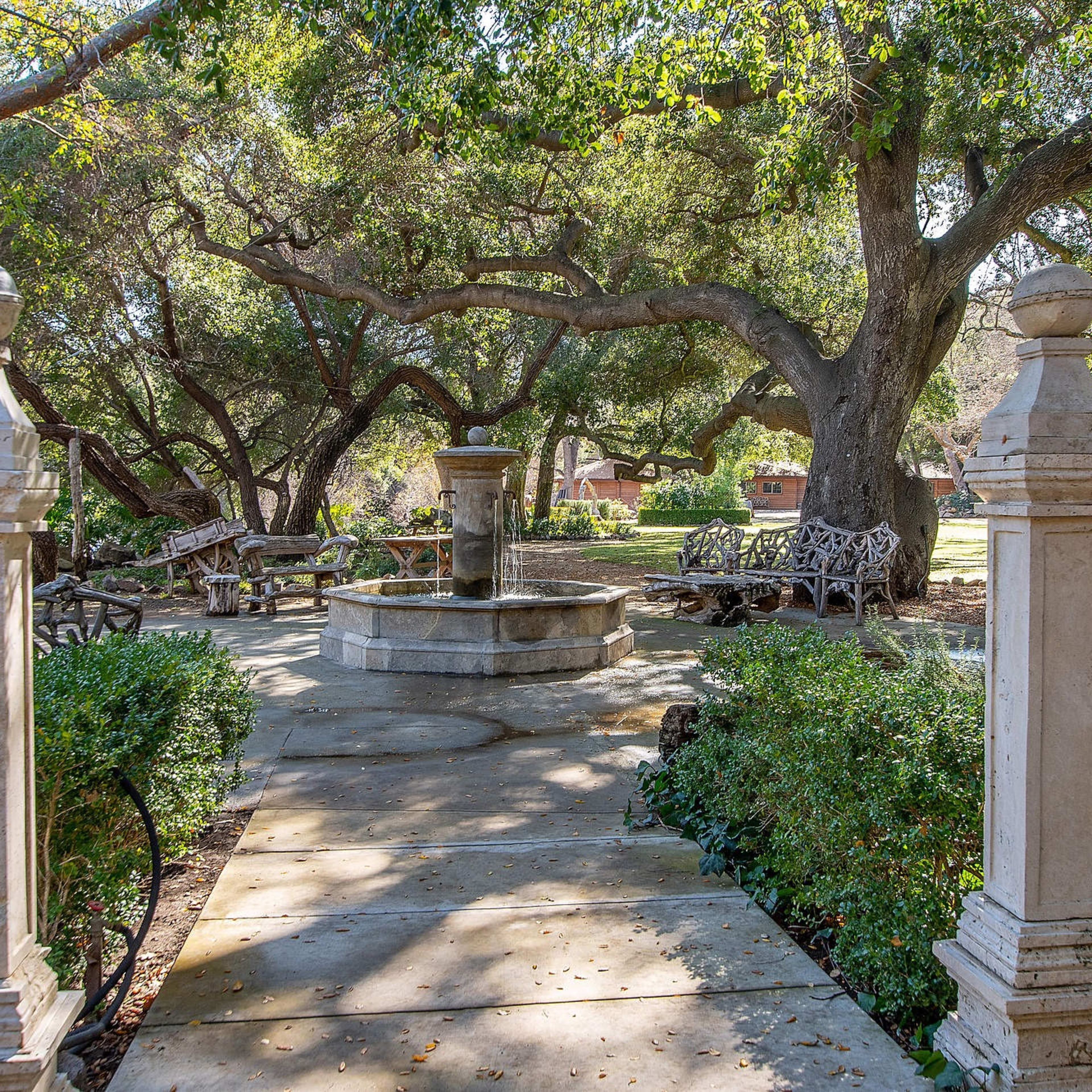 A stone pathway leads to a central fountain surrounded by lush greenery and shaded seating areas beneath large trees.