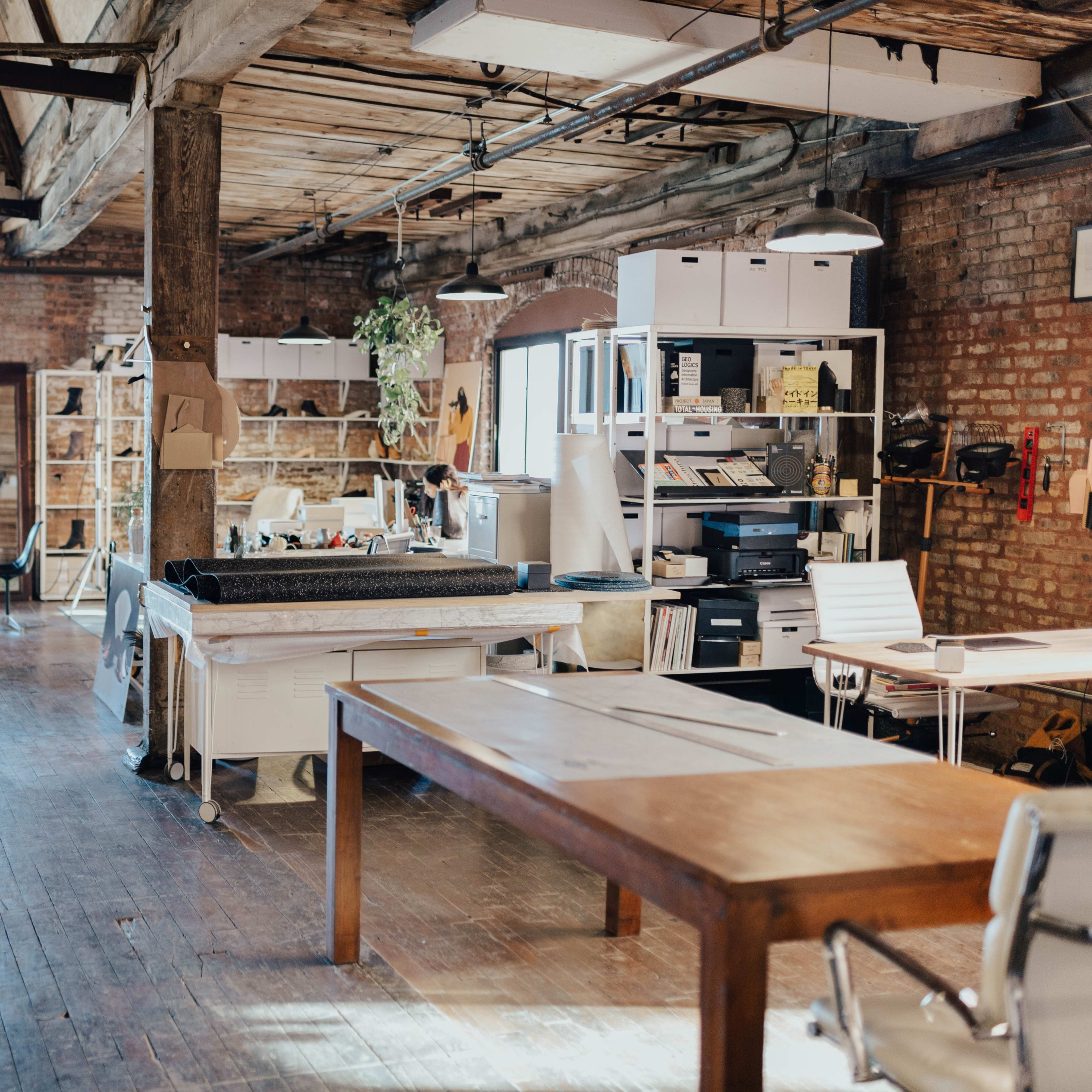 An open-plan office space featuring wooden beams, brick walls, multiple workstations, and a large table in the center.