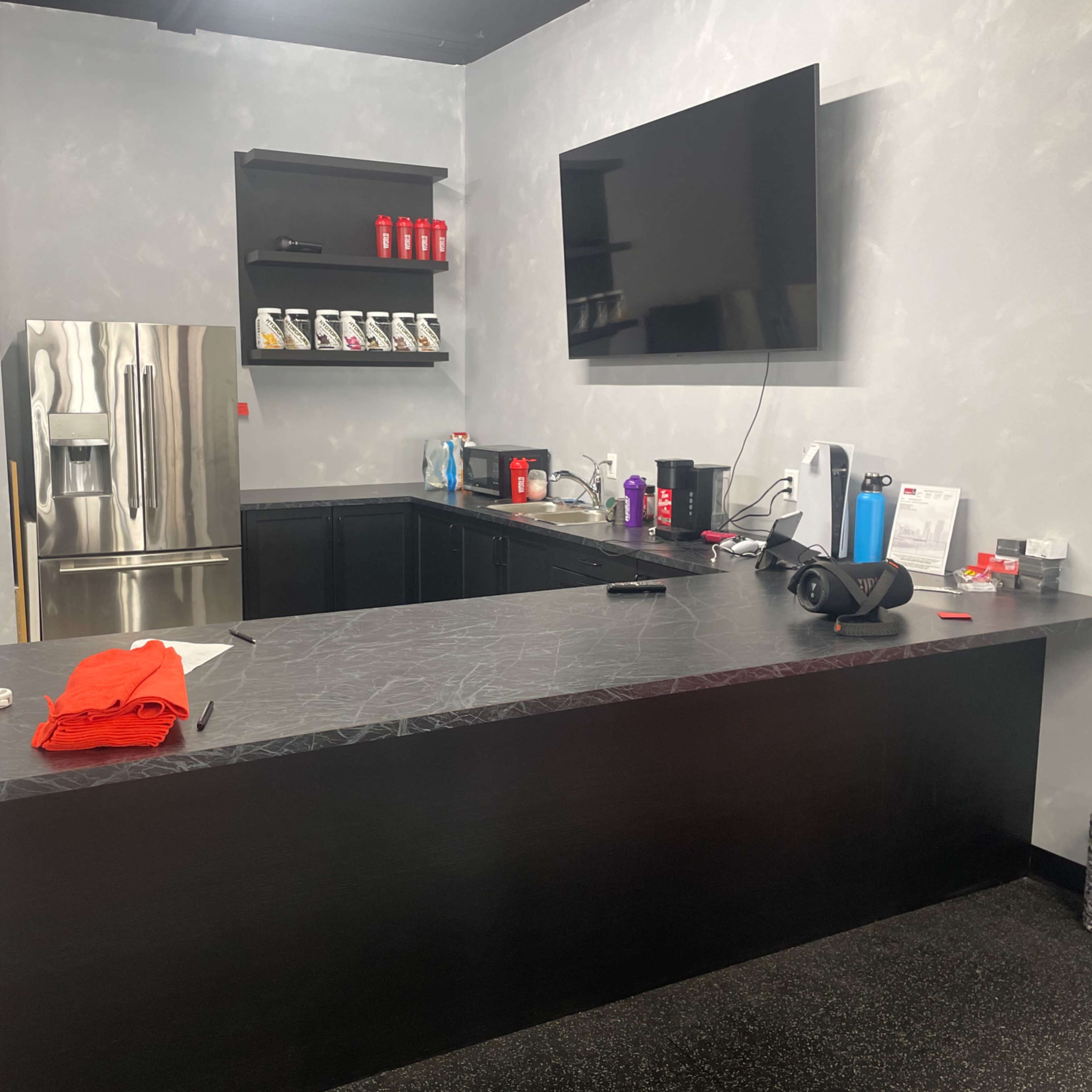 A modern, minimalist kitchenette features a large black countertop, a stainless steel refrigerator, and various appliances arranged on the counter.