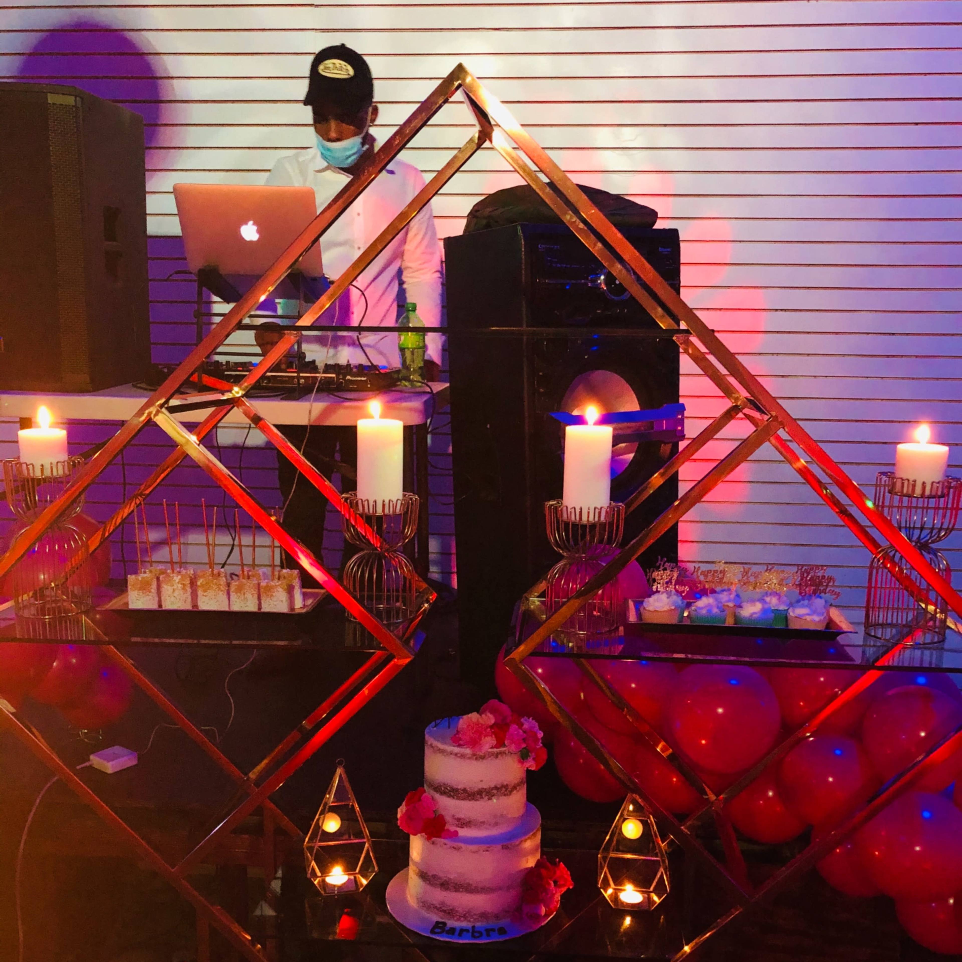 A decorated dessert table with a multi-tiered birthday cake, candles, and assorted treats, set against a backdrop featuring a DJ with a laptop and speaker.
