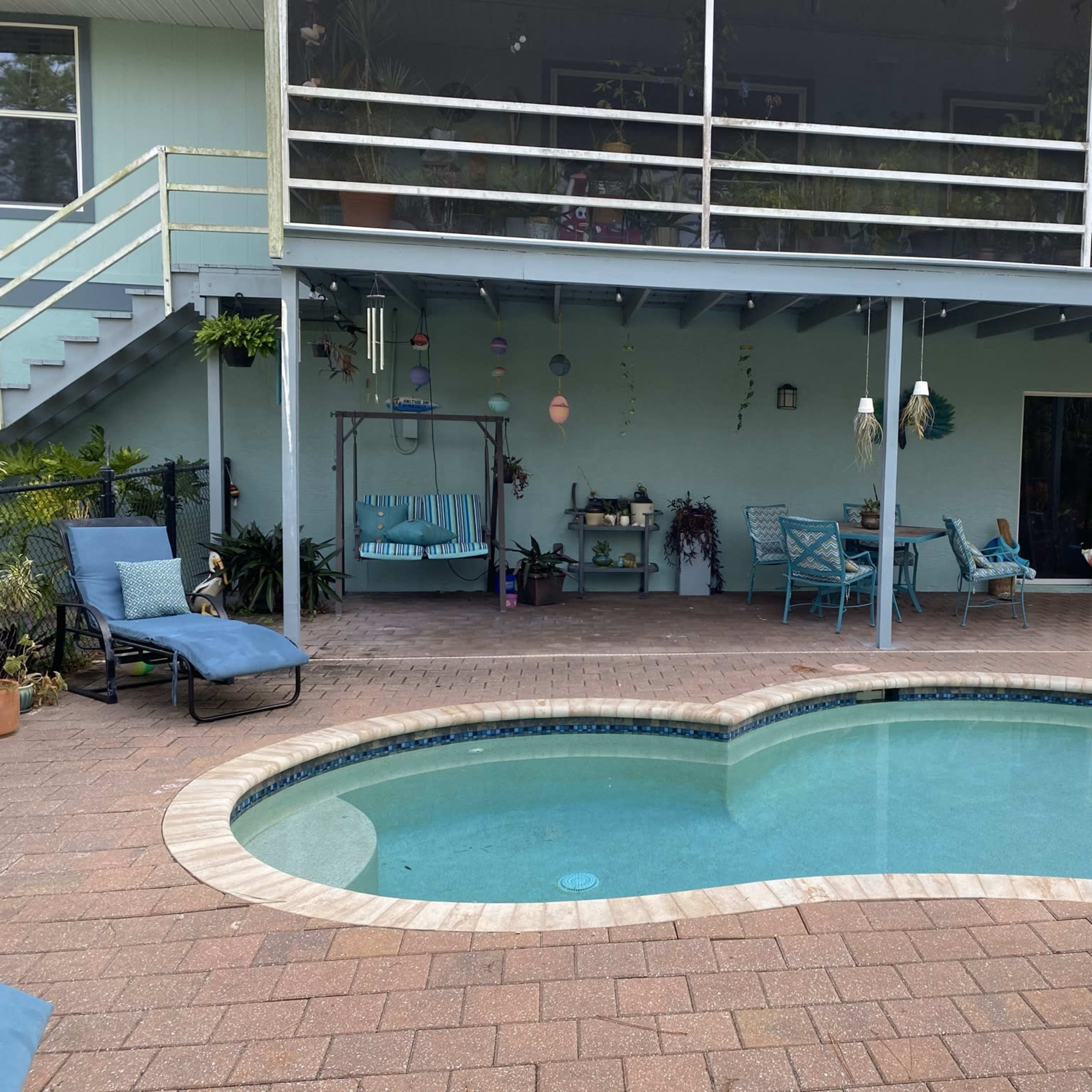 A swimming pool surrounded by a patio with seating areas, a covered deck above, and various plants and decorations.