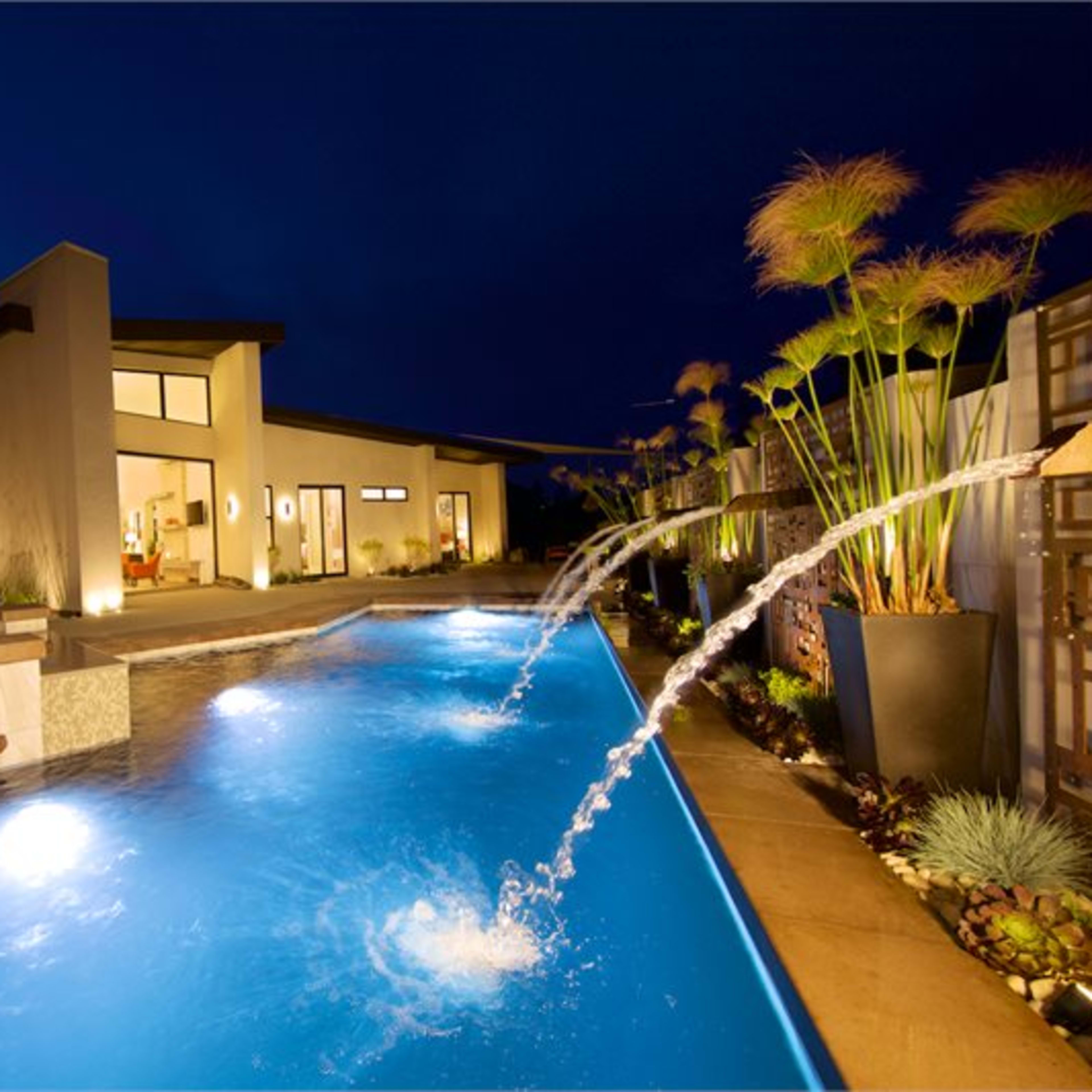A modern home with large windows is illuminated at night, featuring a swimming pool with fountains and landscaping along the edges.