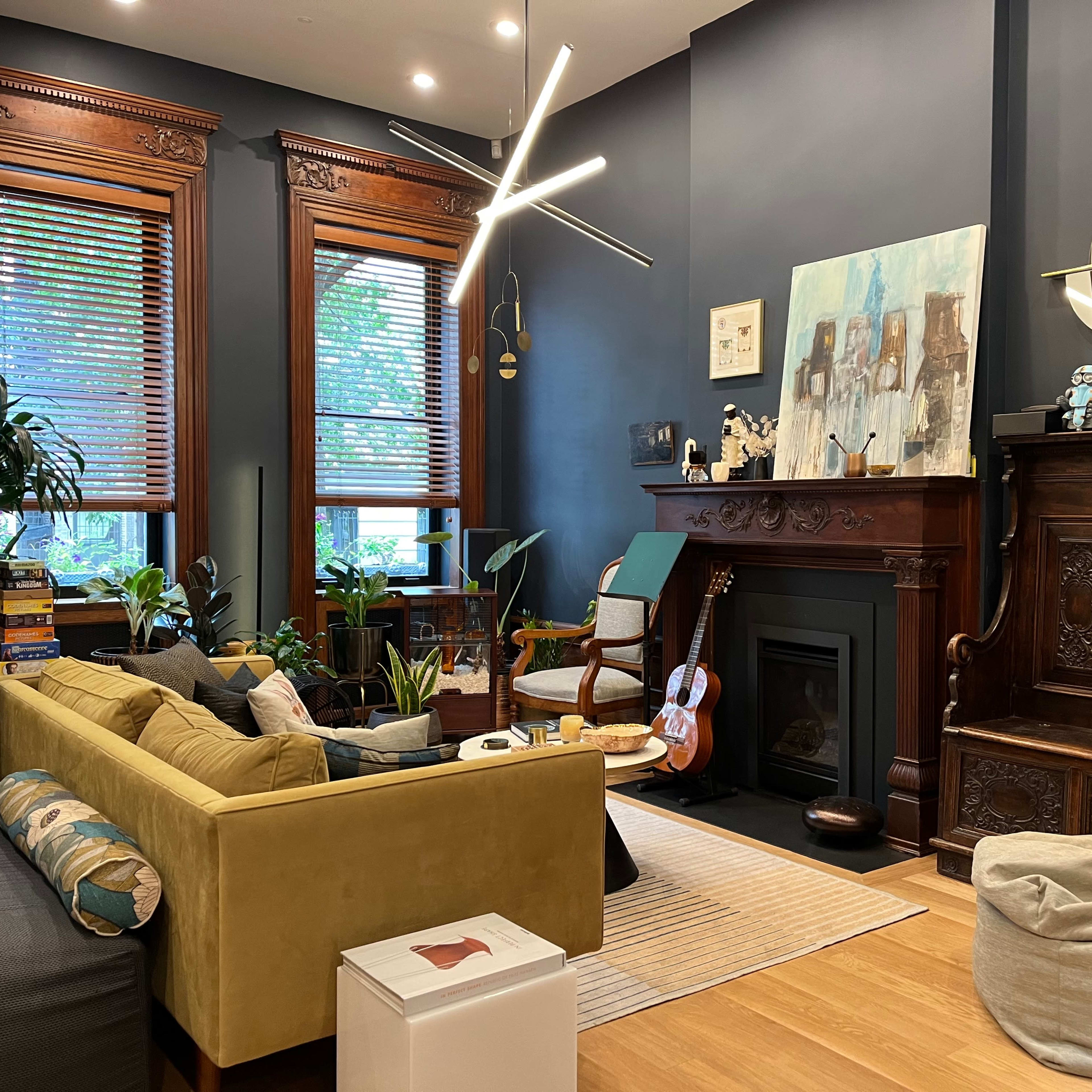 The living room features a green sofa, a guitar, plants, and a decorative wooden fireplace with a wall art piece above it.