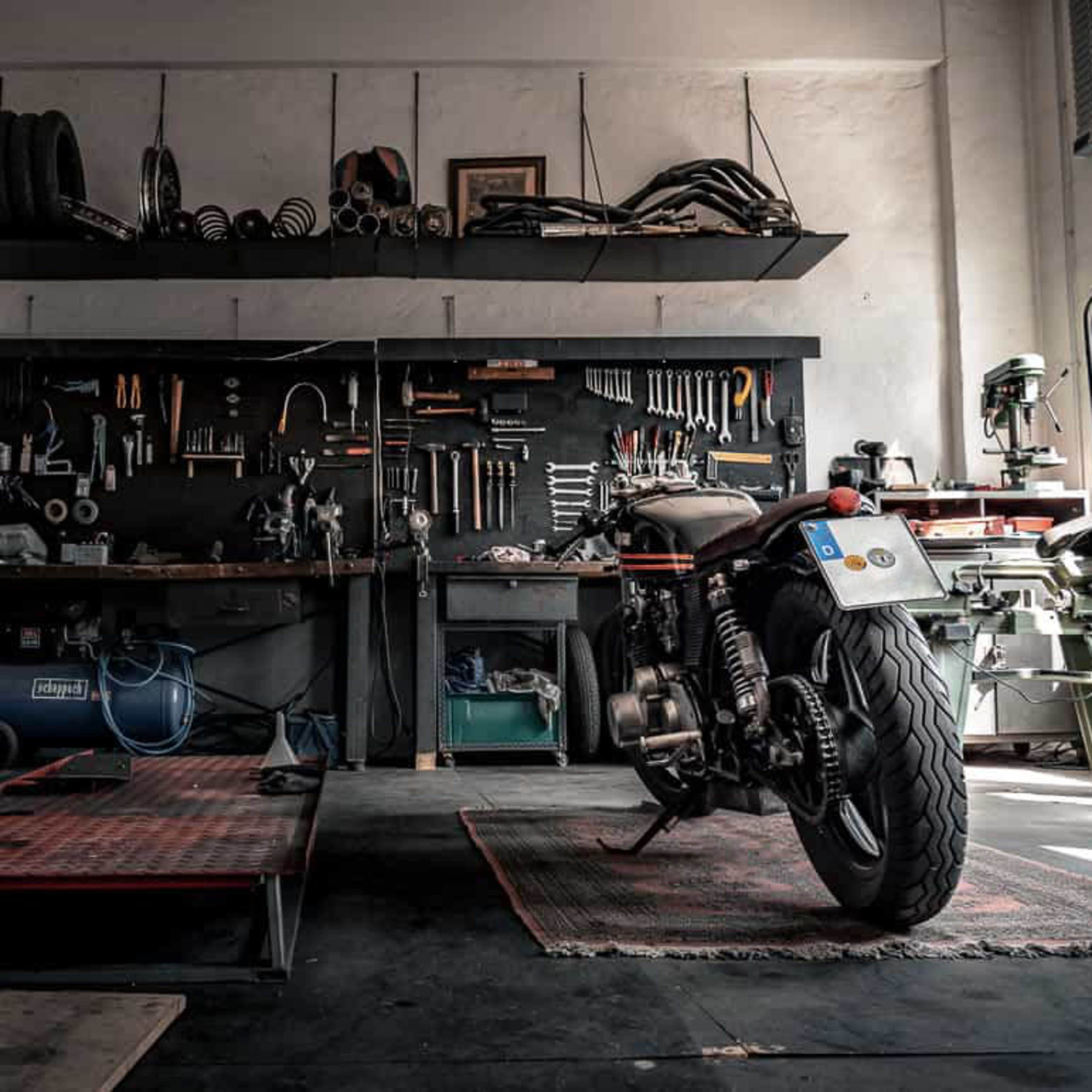 A motorcycle rests on a raised platform in a workshop filled with tools and equipment along the walls.