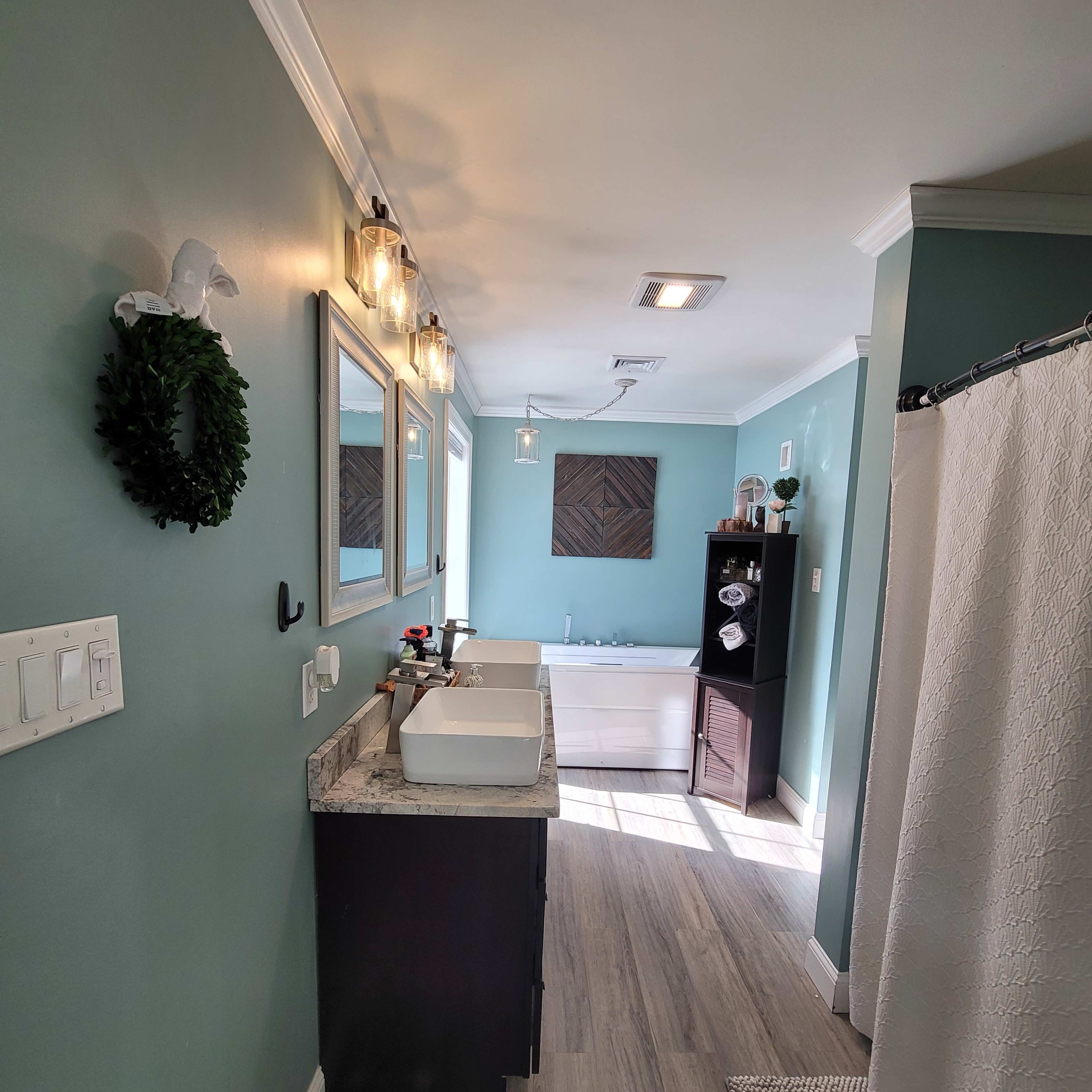 A well-lit bathroom featuring dual sinks with a granite countertop, a tub, and a dark cabinet against a light blue wall.
