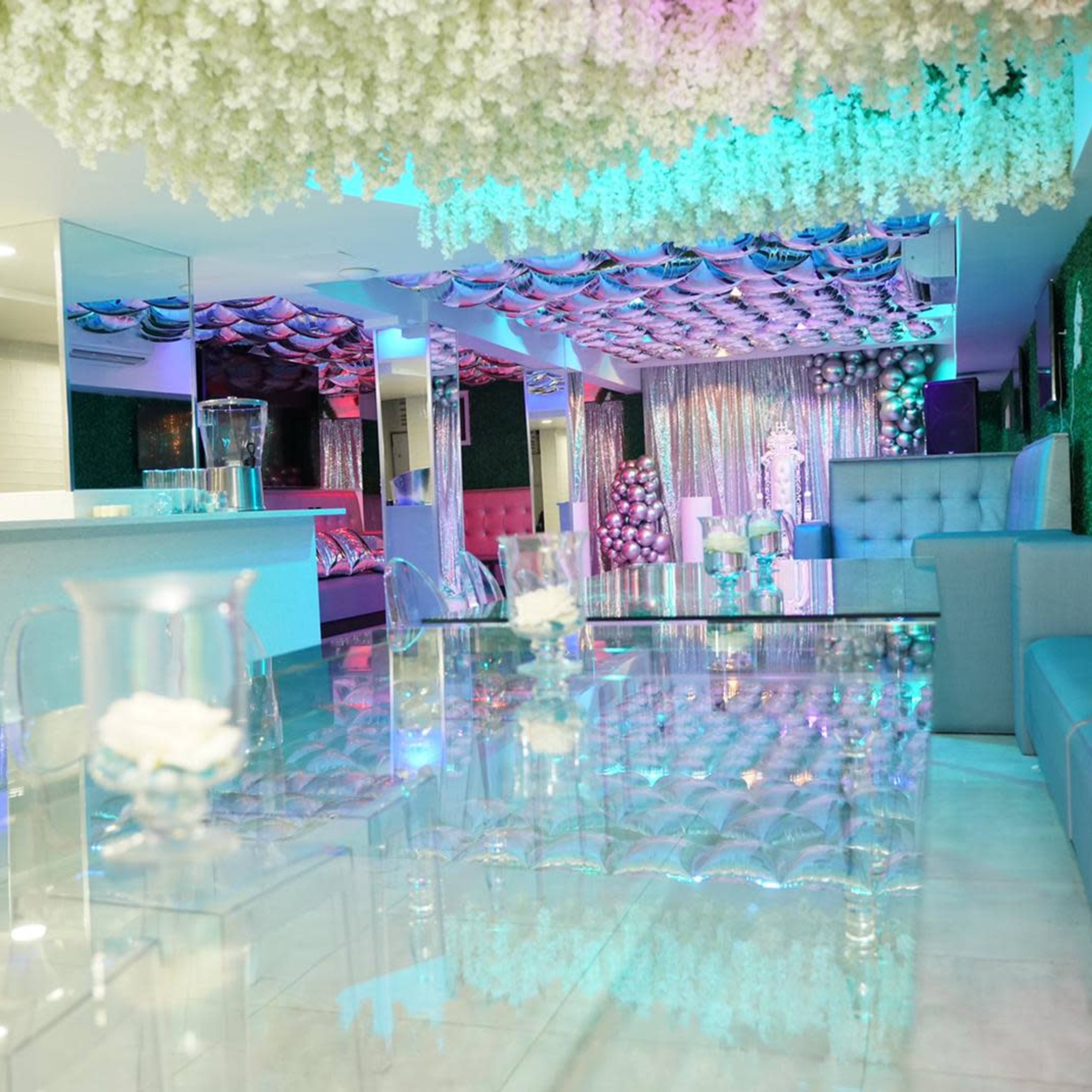 A modern lounge area features transparent furniture, floral decorations overhead, and colorful lighting accents.