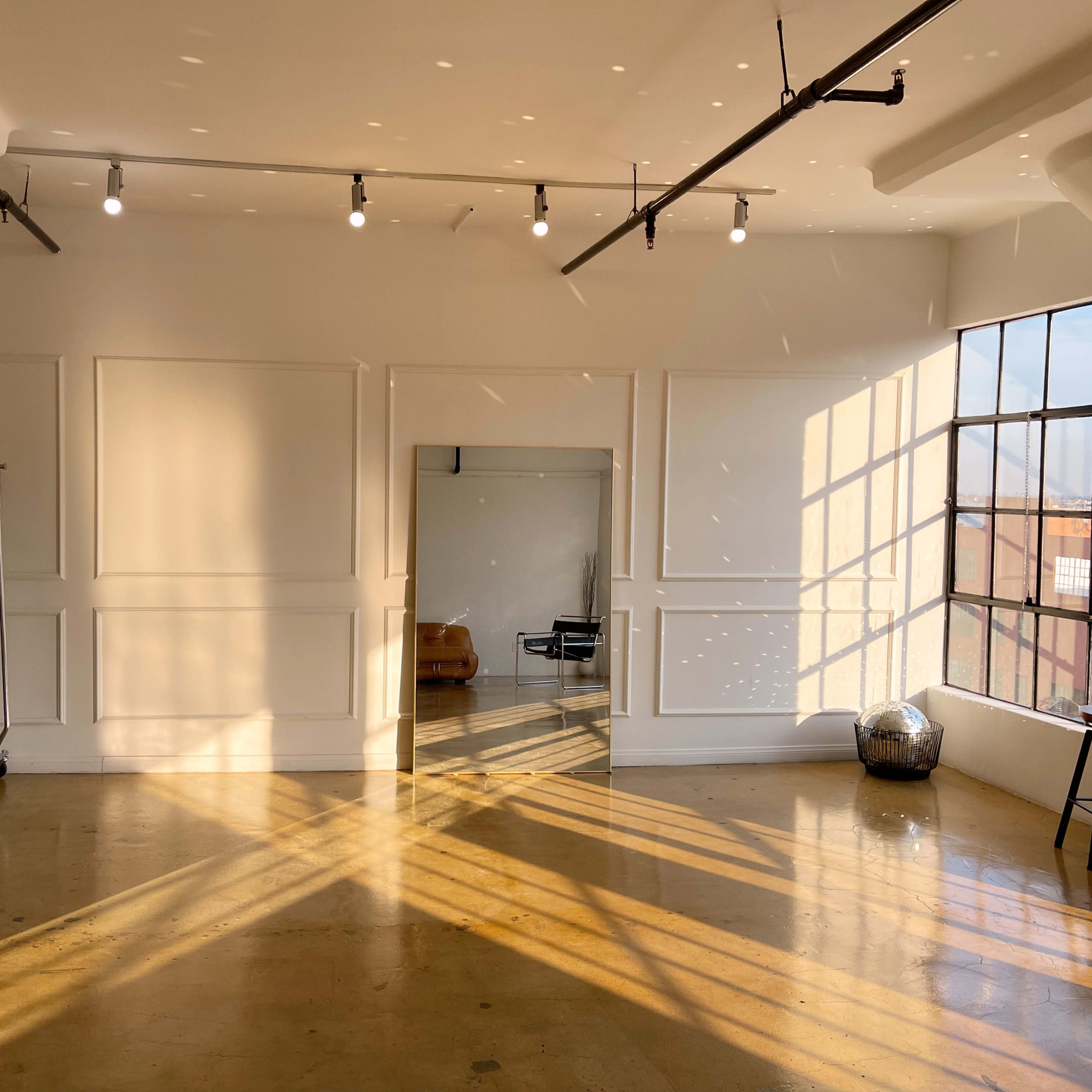 A bright room features a large window, a mirror, a stool, and a clothes rack, with sunlight casting shadows on the polished floor.