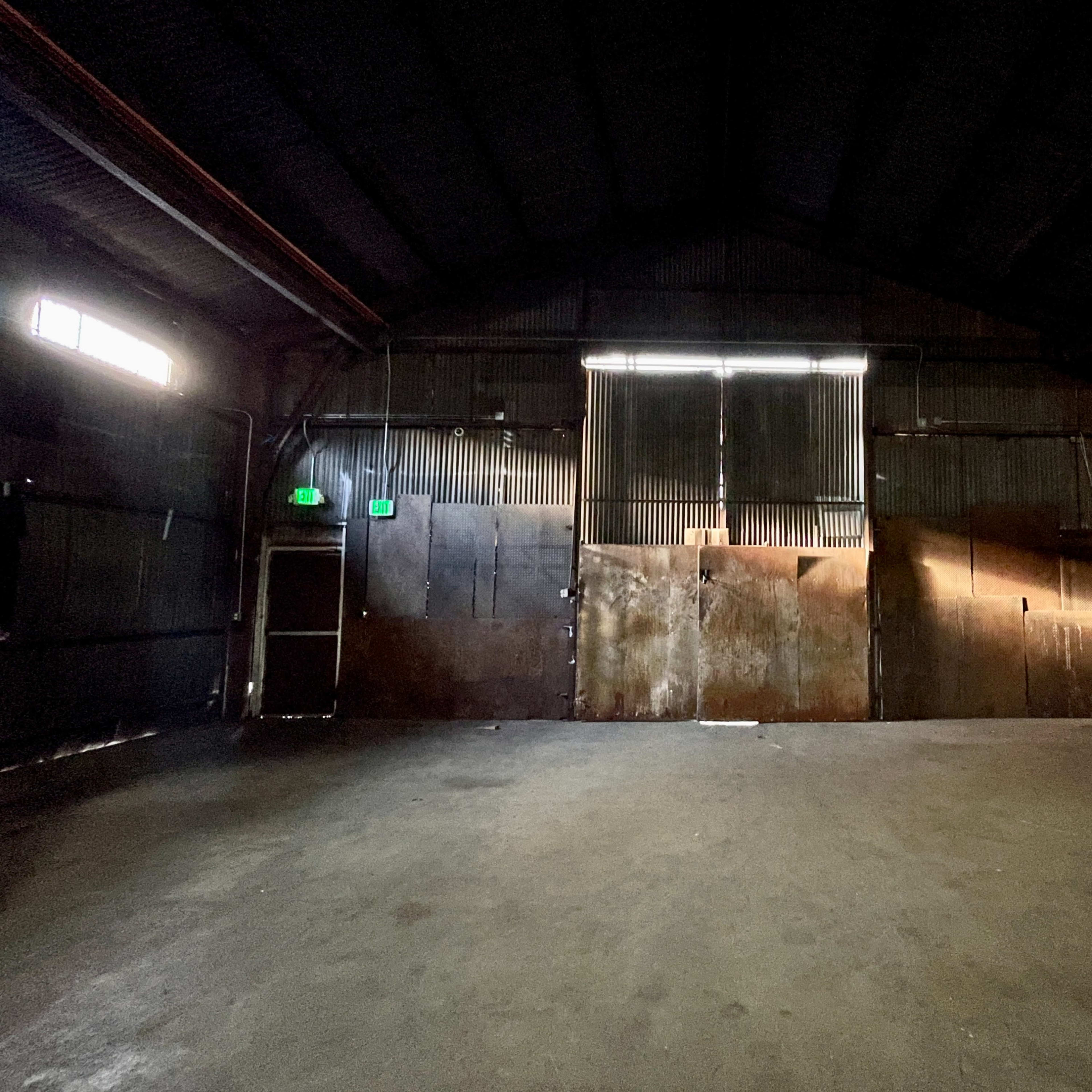 The interior of a large, empty warehouse with metal walls and dim light filtering through partially open doors.