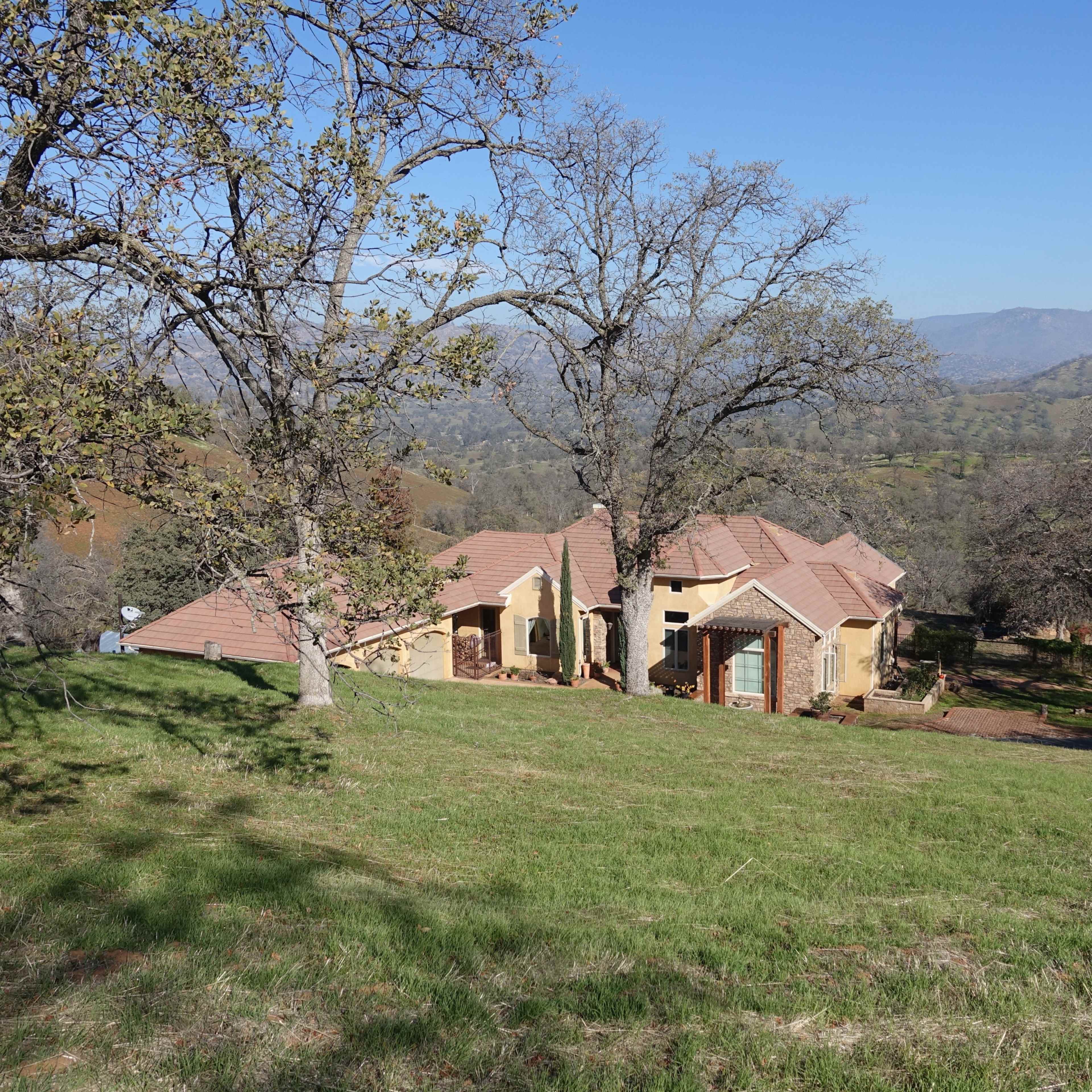 A large, single-story house with a tiled roof is situated on a grassy hillside, surrounded by trees and overlooking a valley.