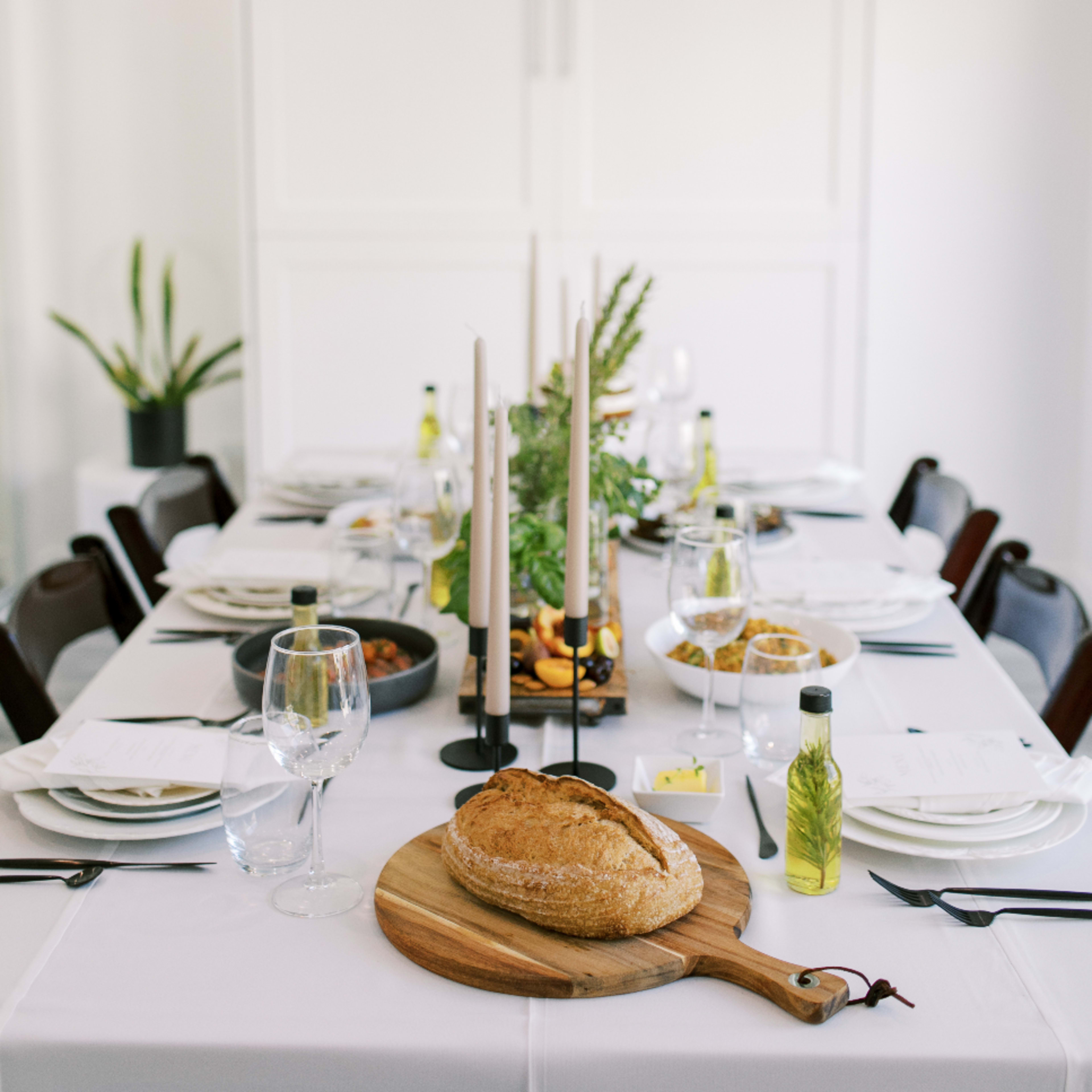 A dining table is set with white tablecloths, various dishes, a loaf of bread on a wooden board, and candles.