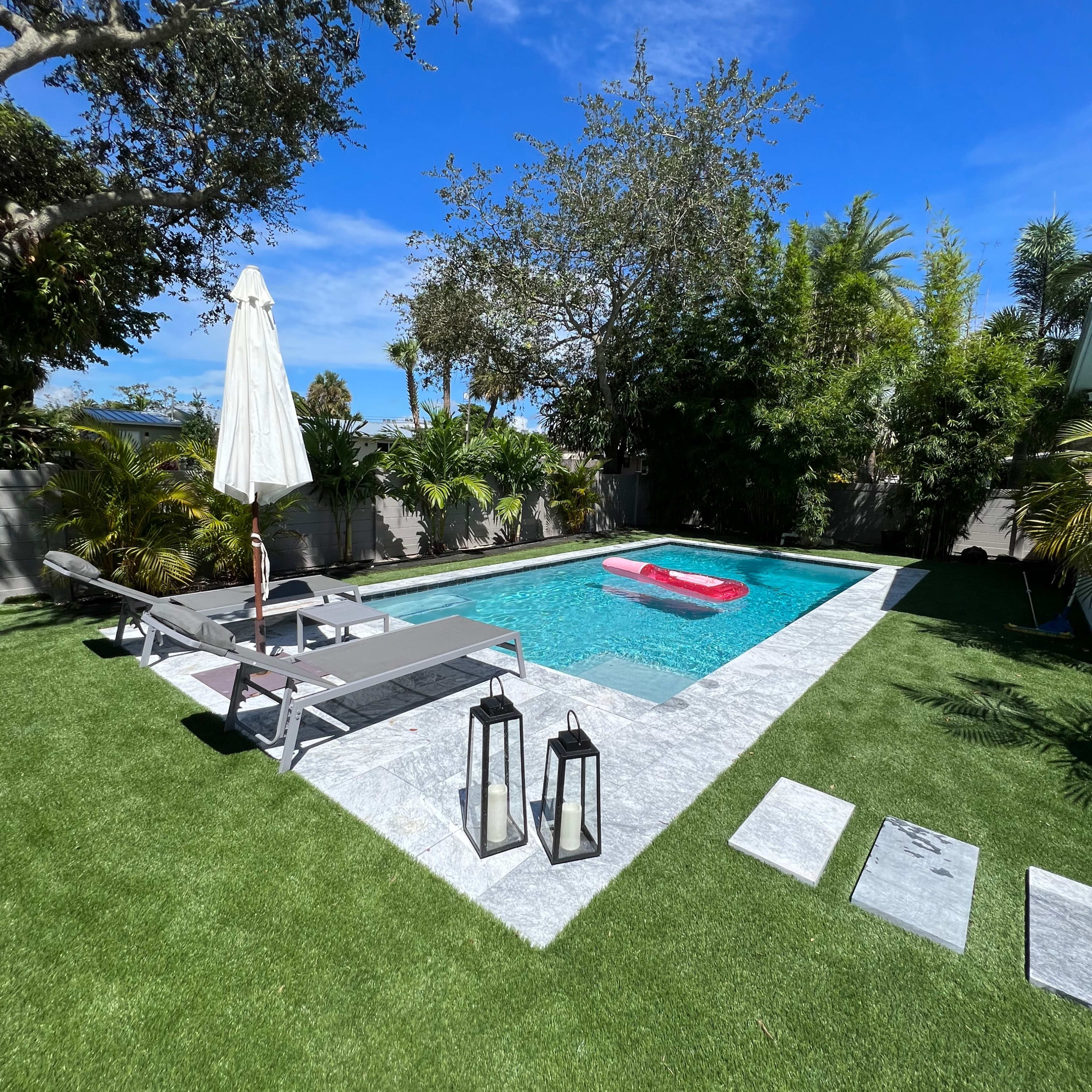 A swimming pool with a lounge area, surrounded by greenery, includes a white umbrella, lounge chairs, and lanterns on a stone path.