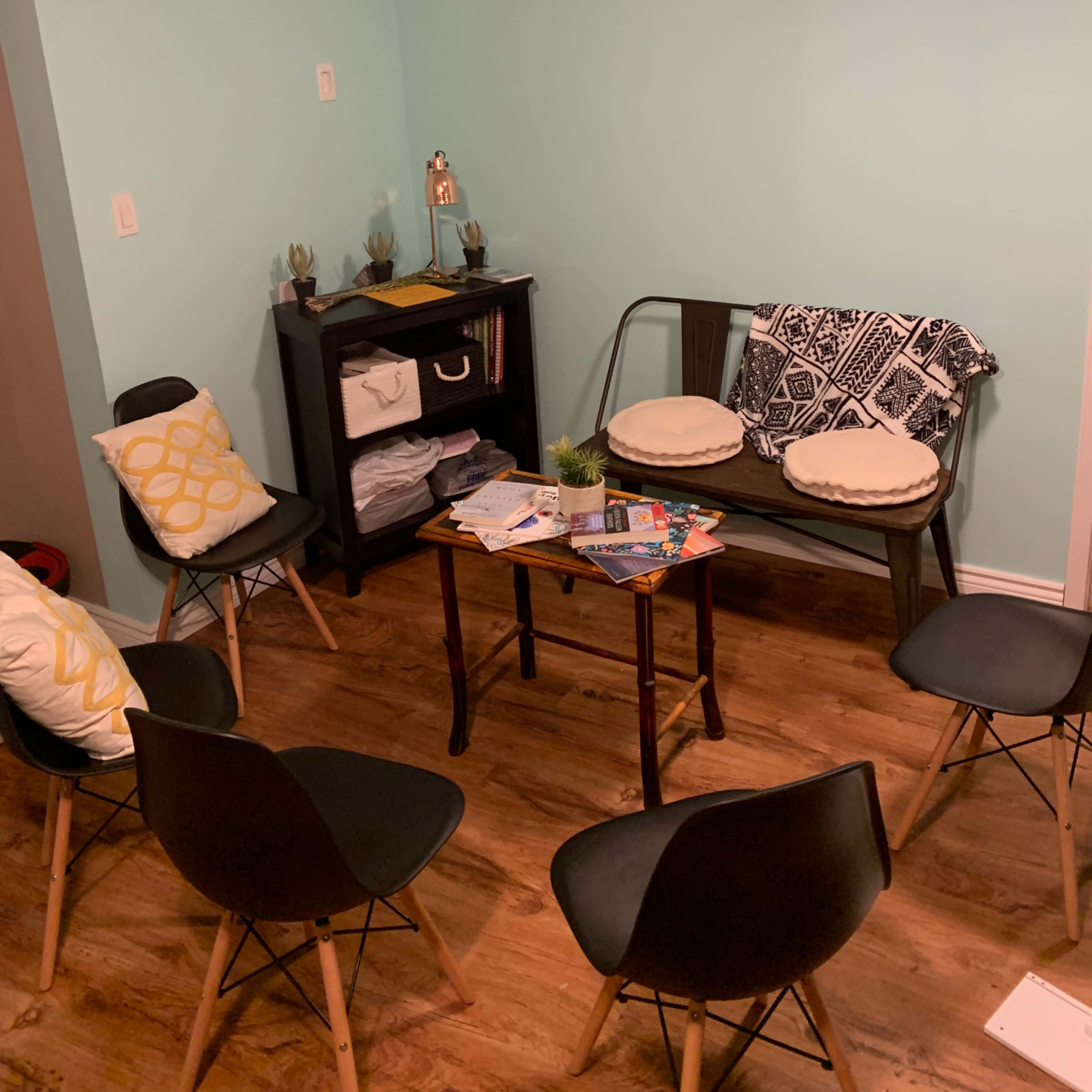 A small room with a seating area arranged around a table, featuring a bench and several black chairs, with a bookshelf in the corner.