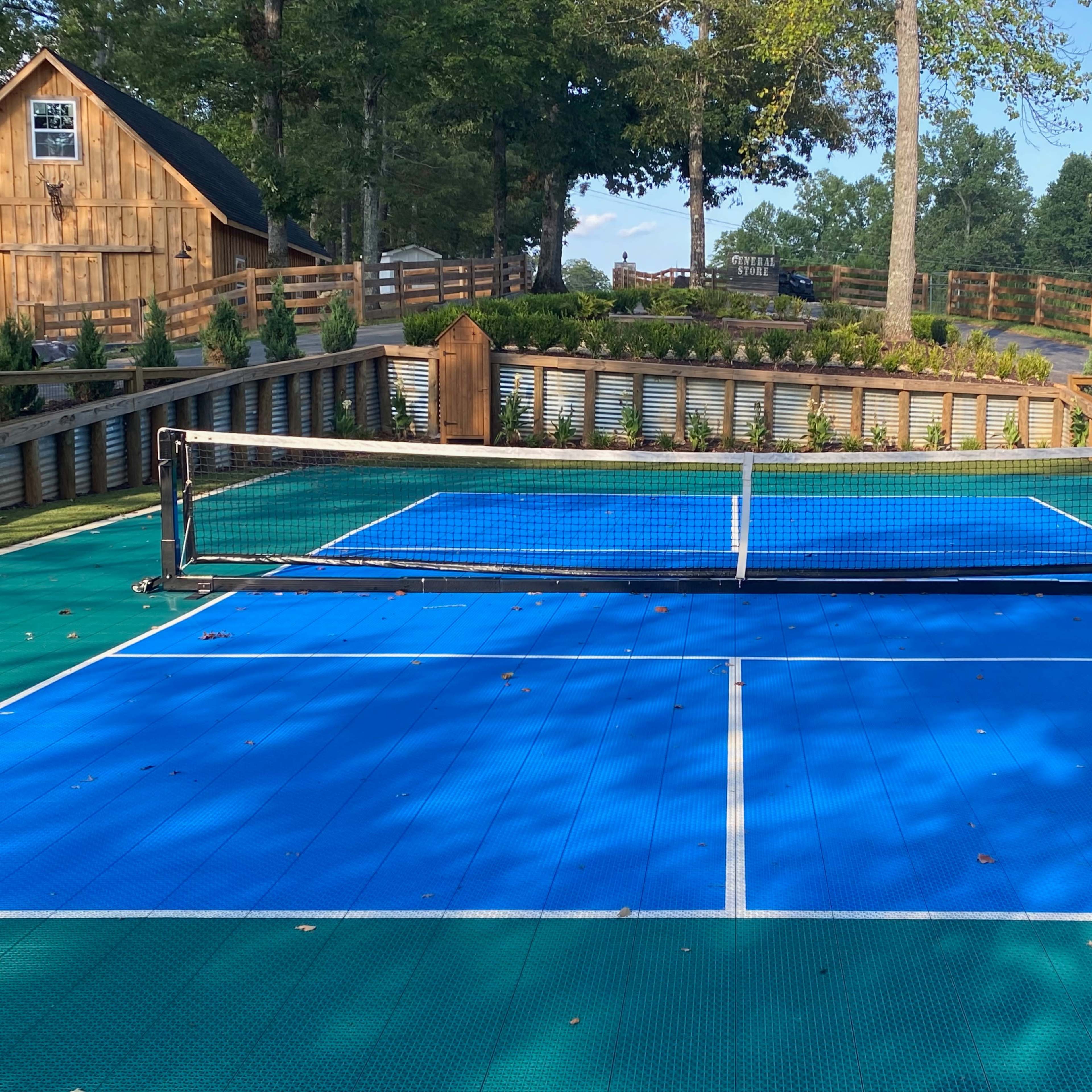 A tennis court with a blue surface and a net, surrounded by trees and a wooden fence.