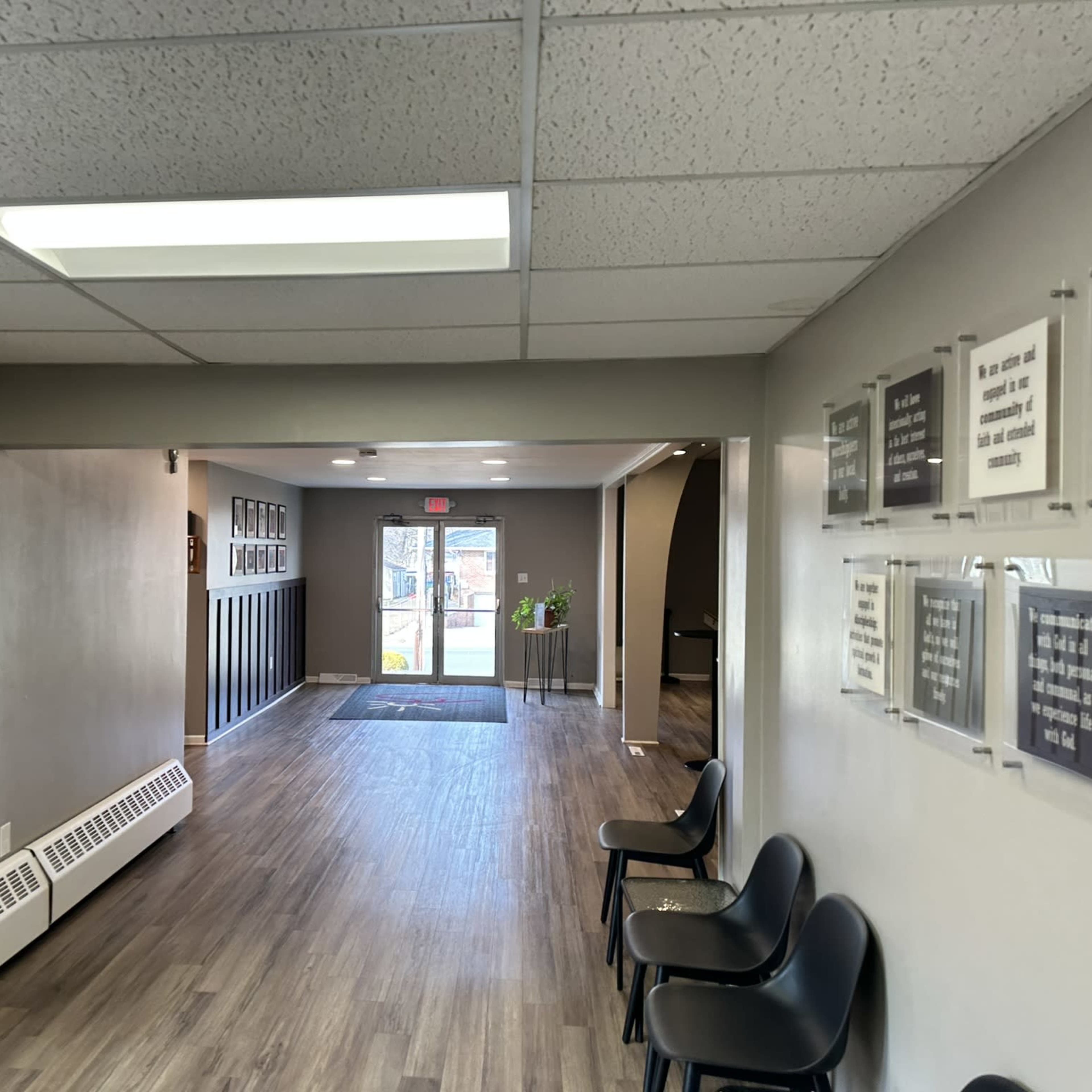 A hallway with a clean floor, leading to a door at the end, flanked by black chairs and framed signs on the walls.