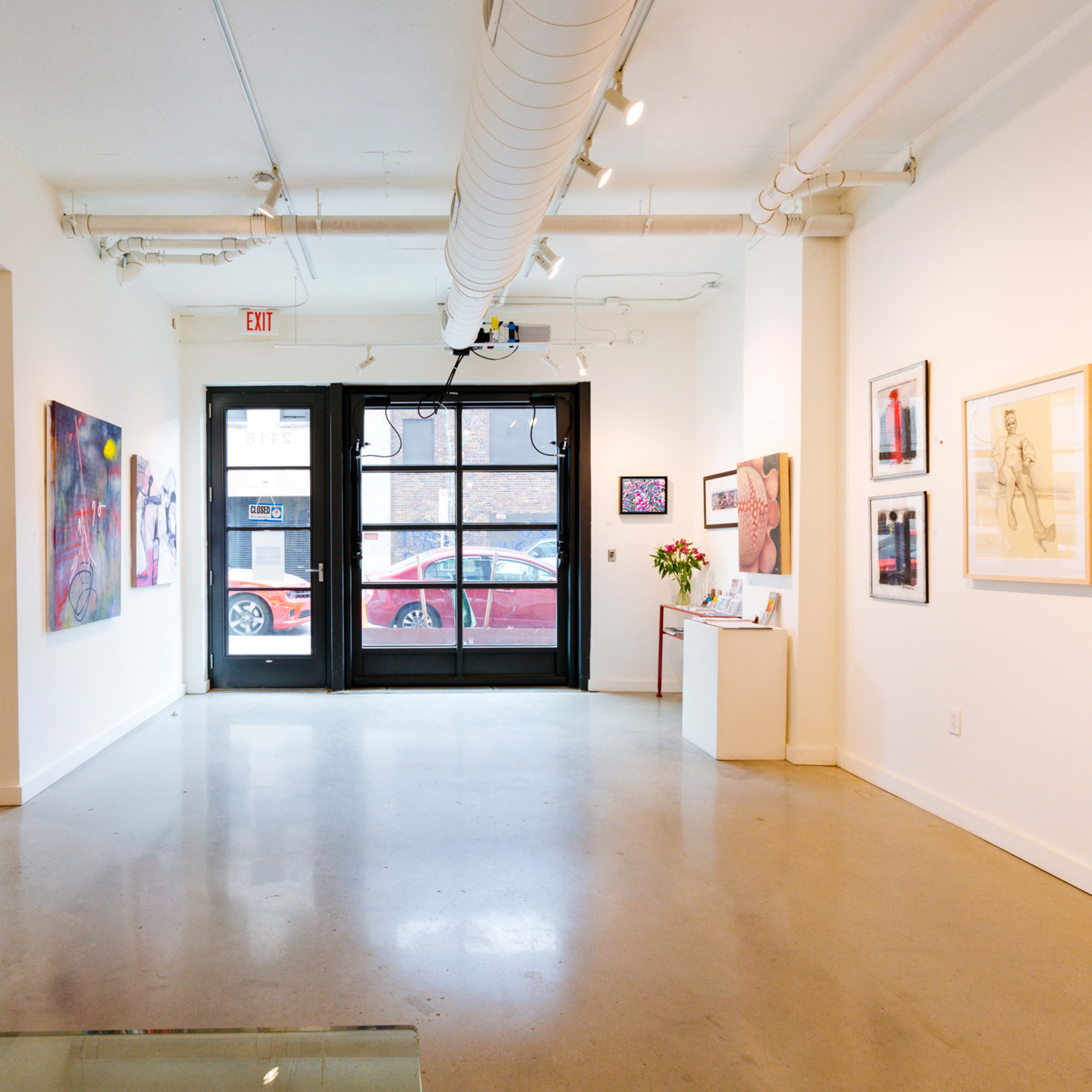 A spacious art gallery with white walls, featuring framed artworks and large glass doors leading to the street outside.