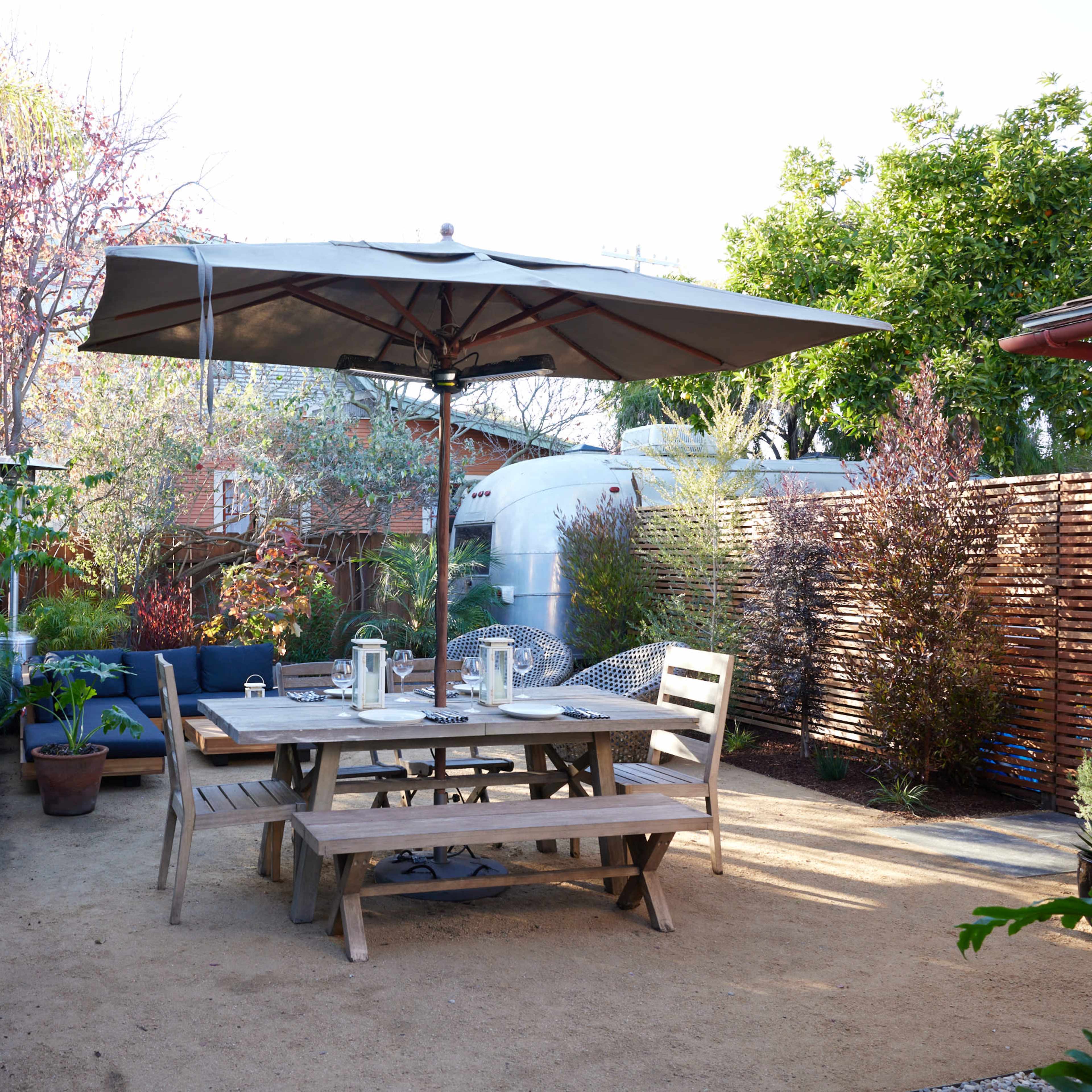 A wooden dining table with chairs sits under a large umbrella in a landscaped outdoor space featuring a grill, plants, and a wooden fence.