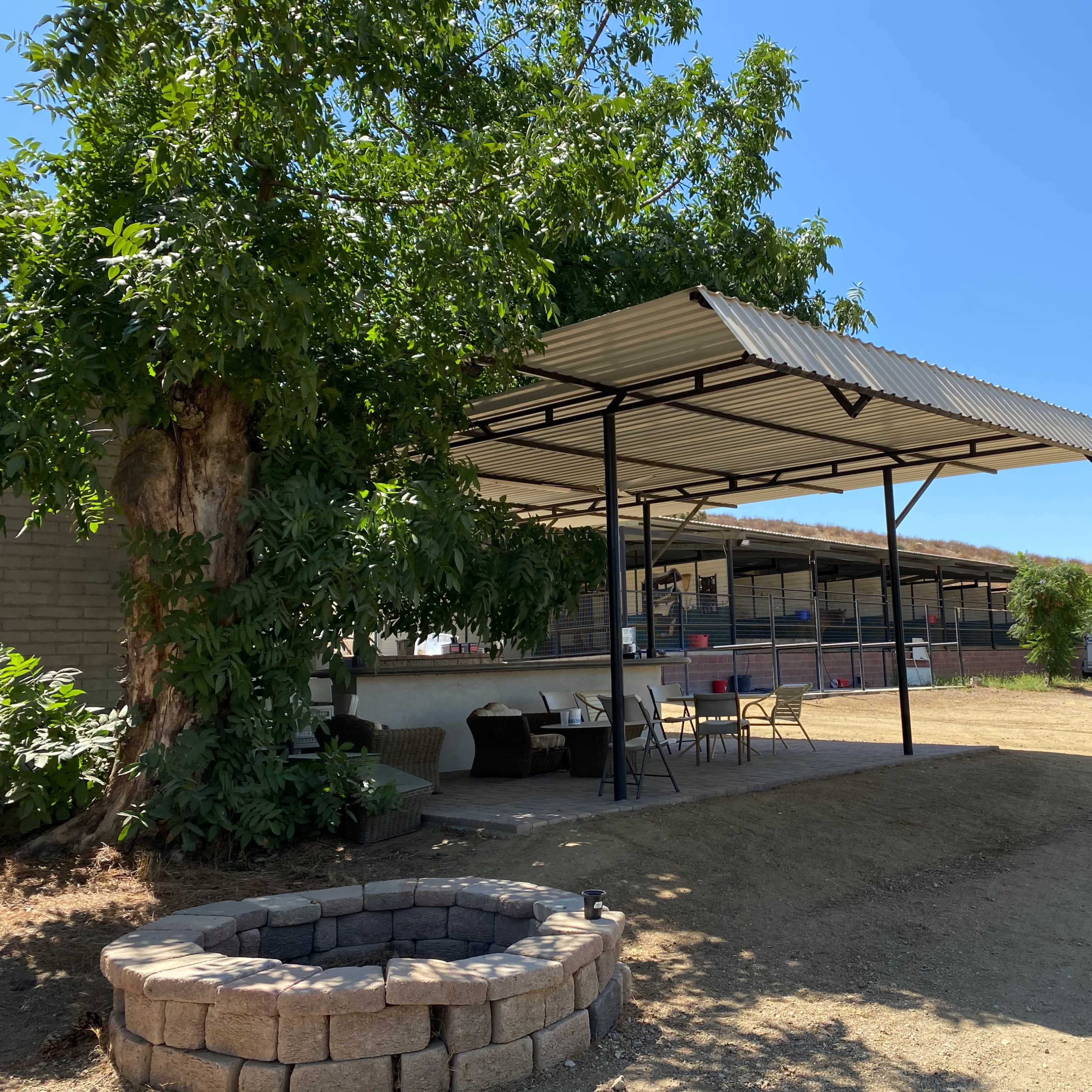 A shaded area with chairs and tables under a roof beside a circular stone fire pit and structures in the background.