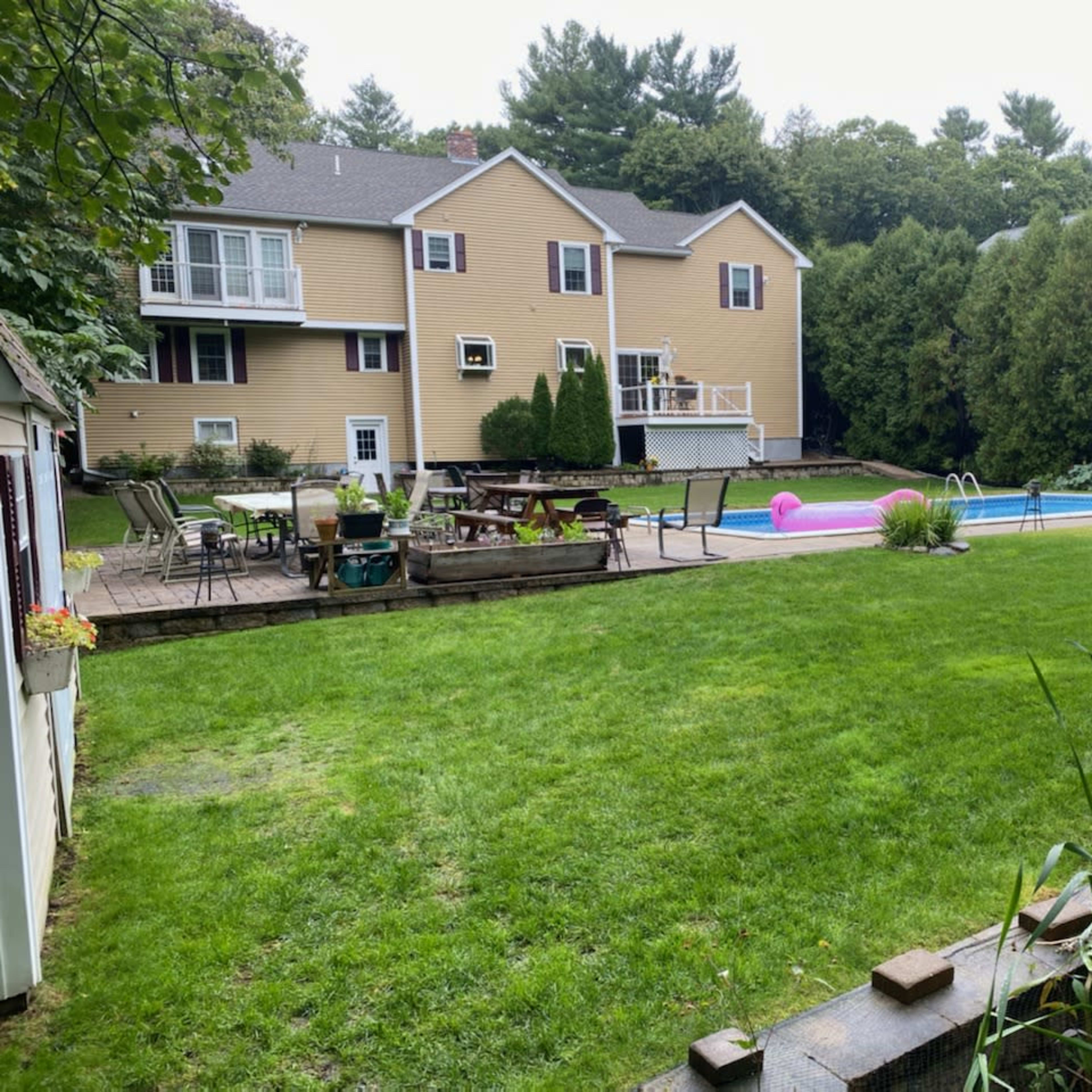 A backyard with a pool, lawn chairs, and a large green lawn in front of a two-story yellow house.