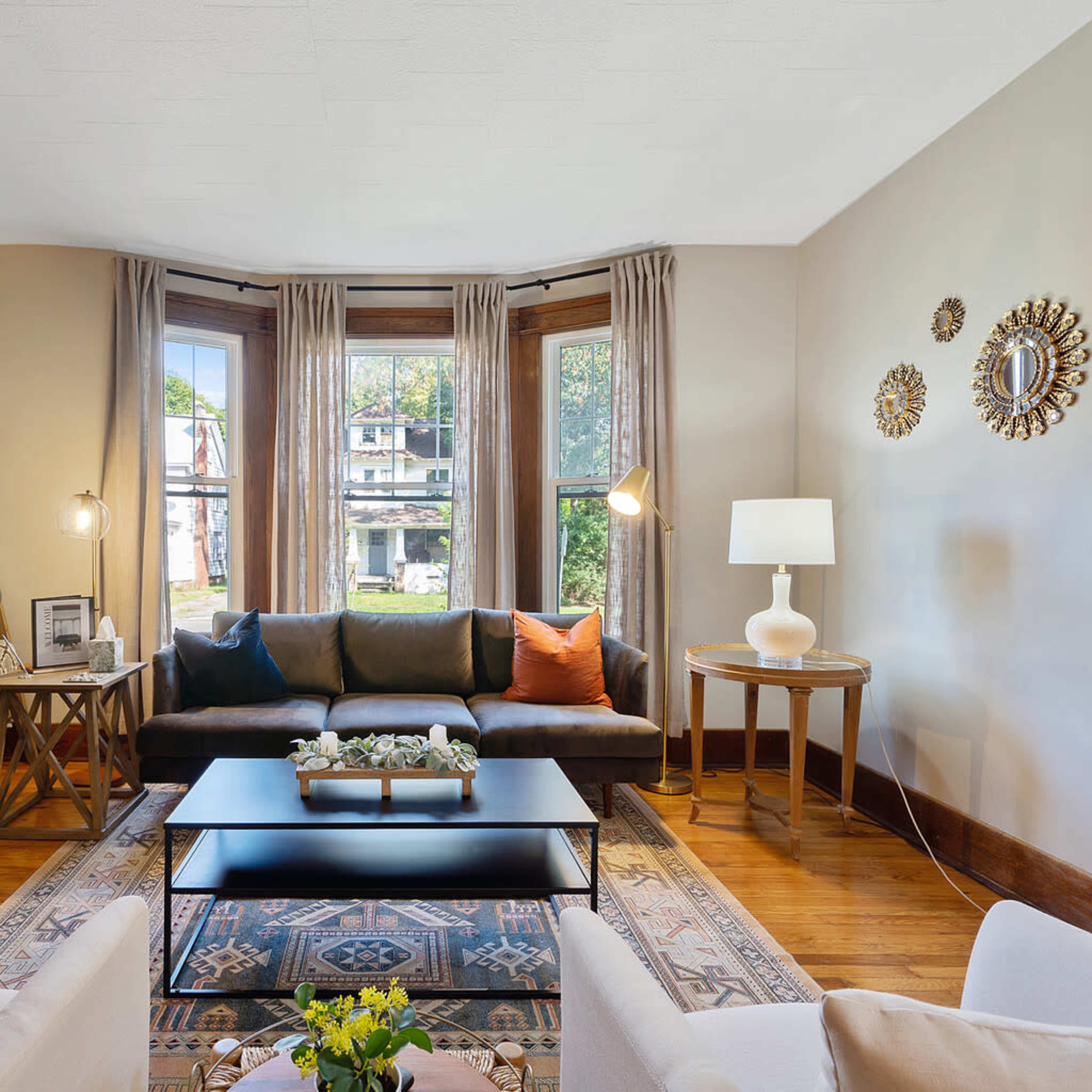 A cozy living room featuring a fireplace, a sectional sofa, large windows with curtains, and a coffee table arranged with decorative items.