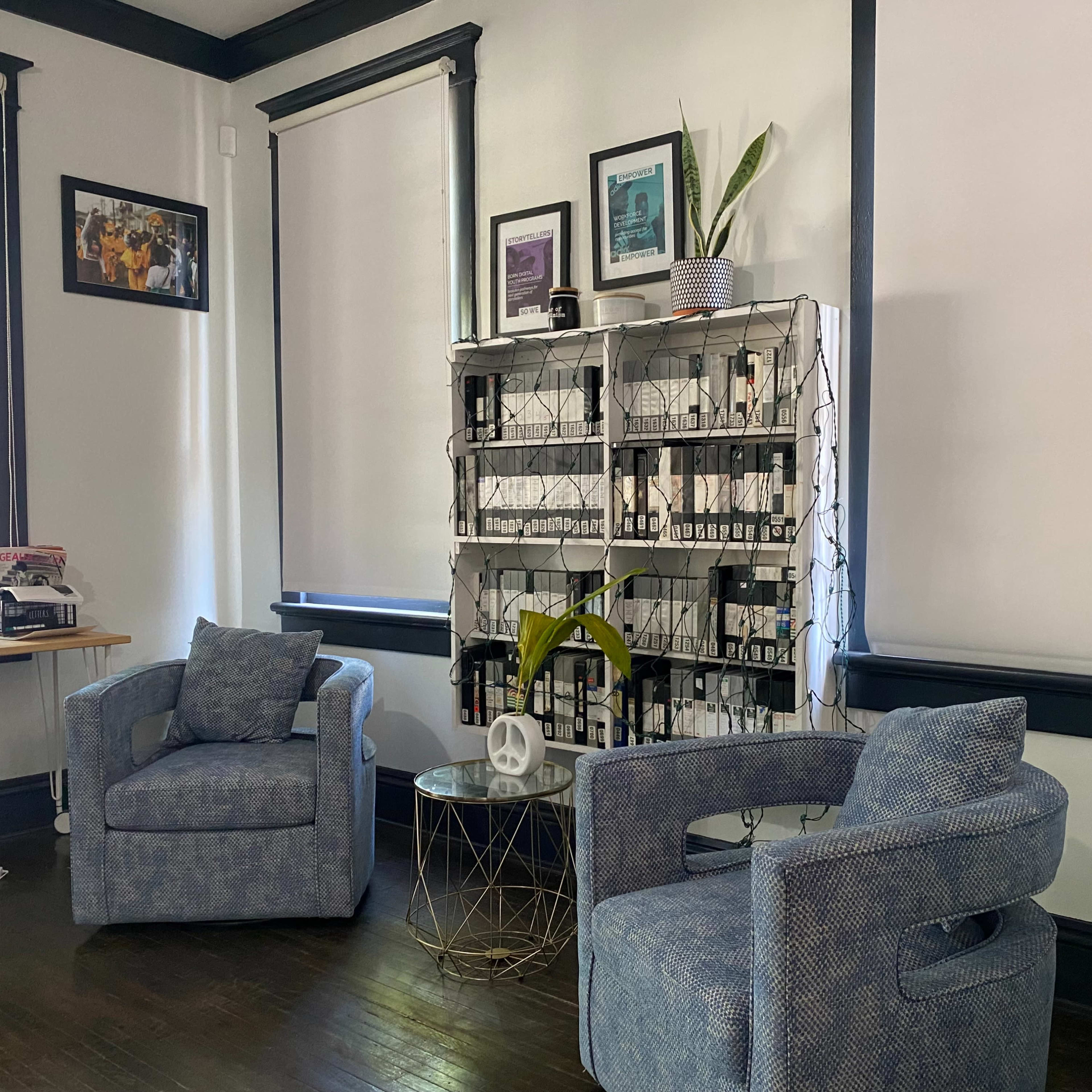 A cozy room with two blue armchairs positioned near a shelf filled with bottles and decorated with plants, alongside a small glass and metal side table.