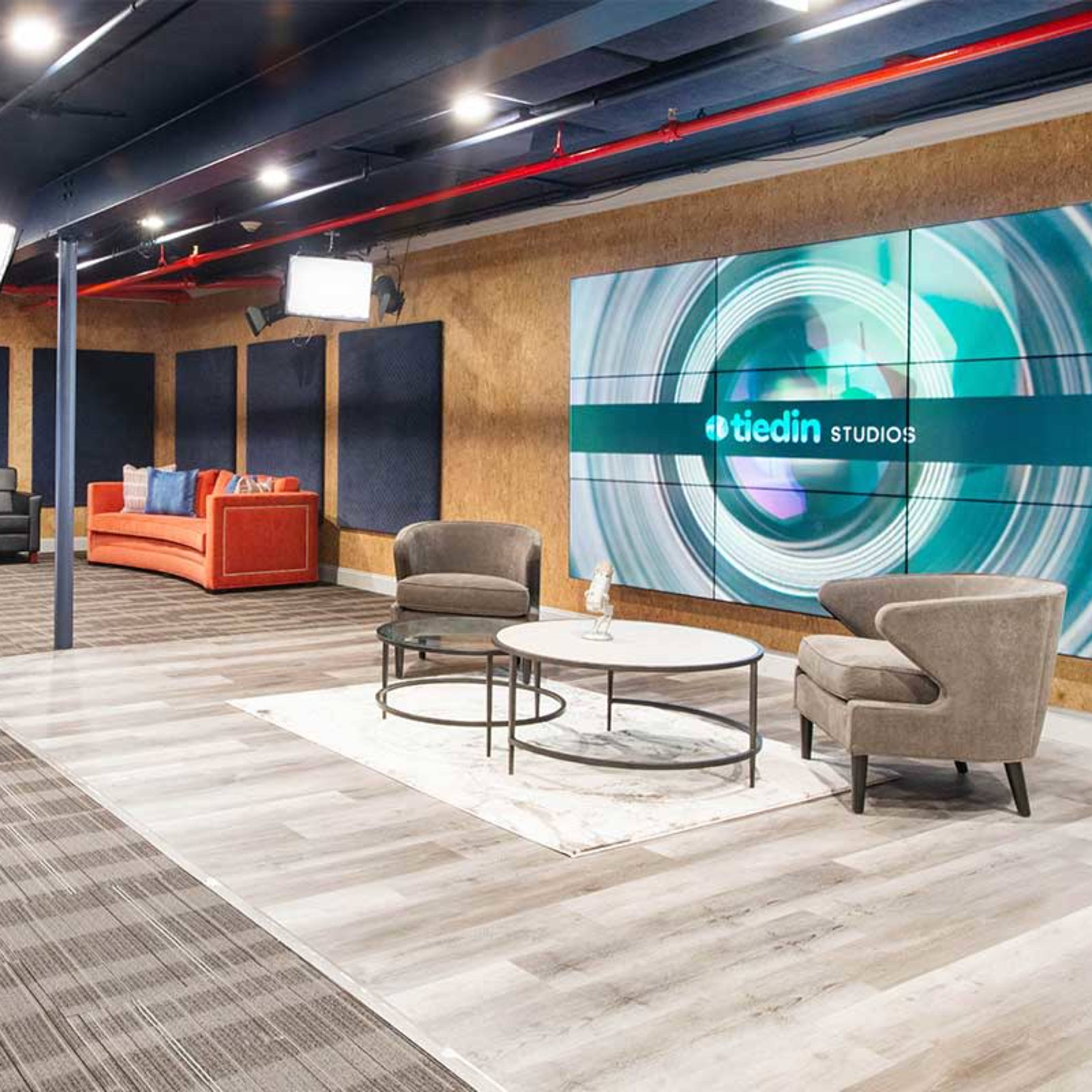 A modern studio setting featuring a beige and blue color scheme, with comfortable seating, professional lighting, and a large screen displaying the logo of a media company.