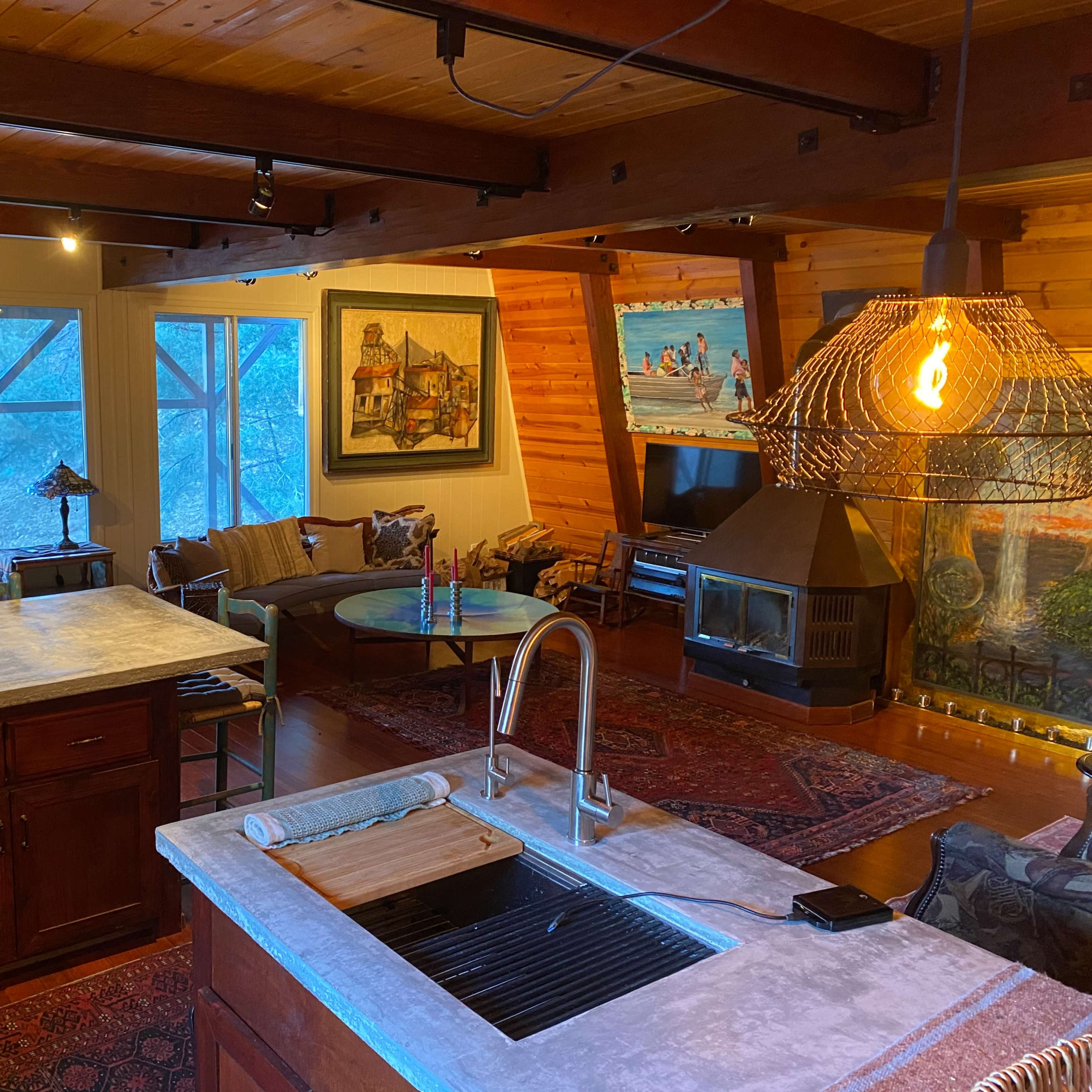 The interior of a cozy, well-lit living space features a kitchen island, a seating area with large windows, and several colorful paintings on the walls.