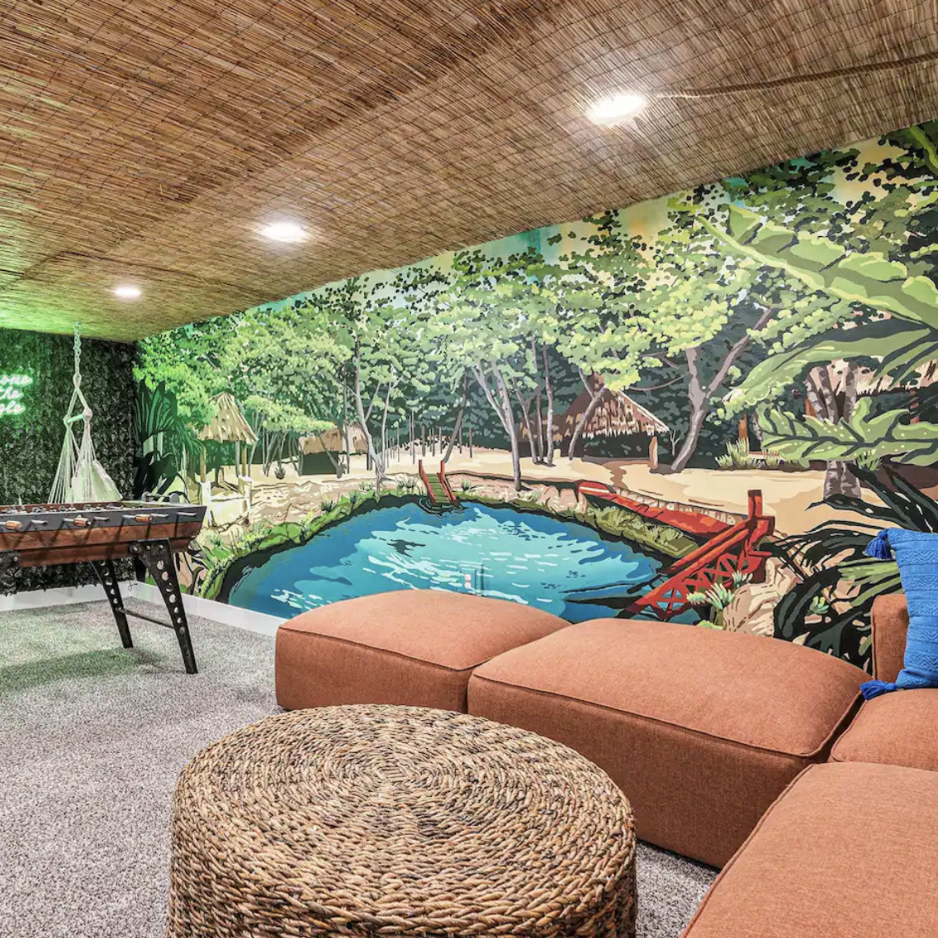 A cozy basement area featuring a foosball table, a large mural of a tropical scene, and stylish seating arrangements.