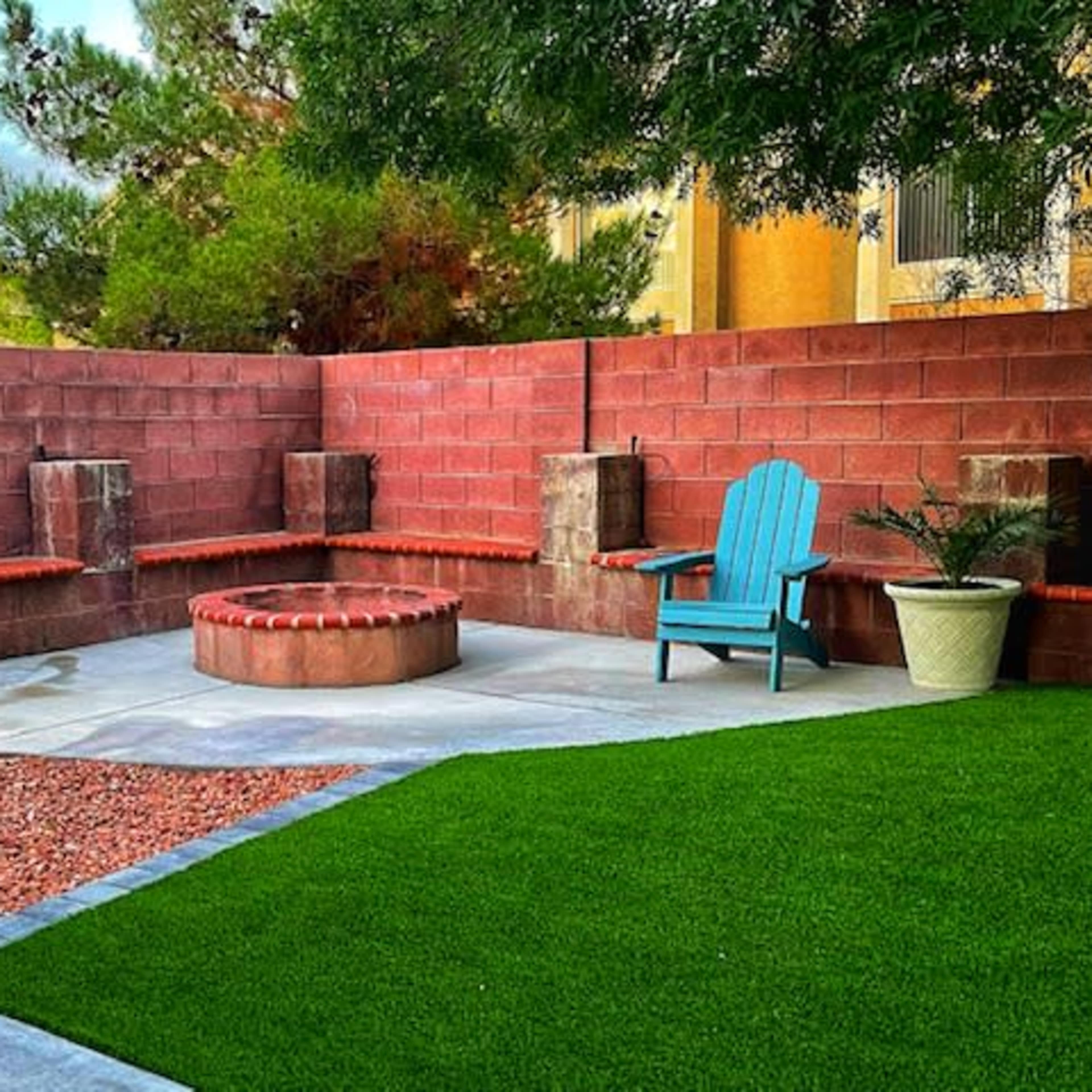 A backyard patio featuring a fire pit surrounded by two stone benches, an adirondack chair, and landscaped areas with grass and gravel.