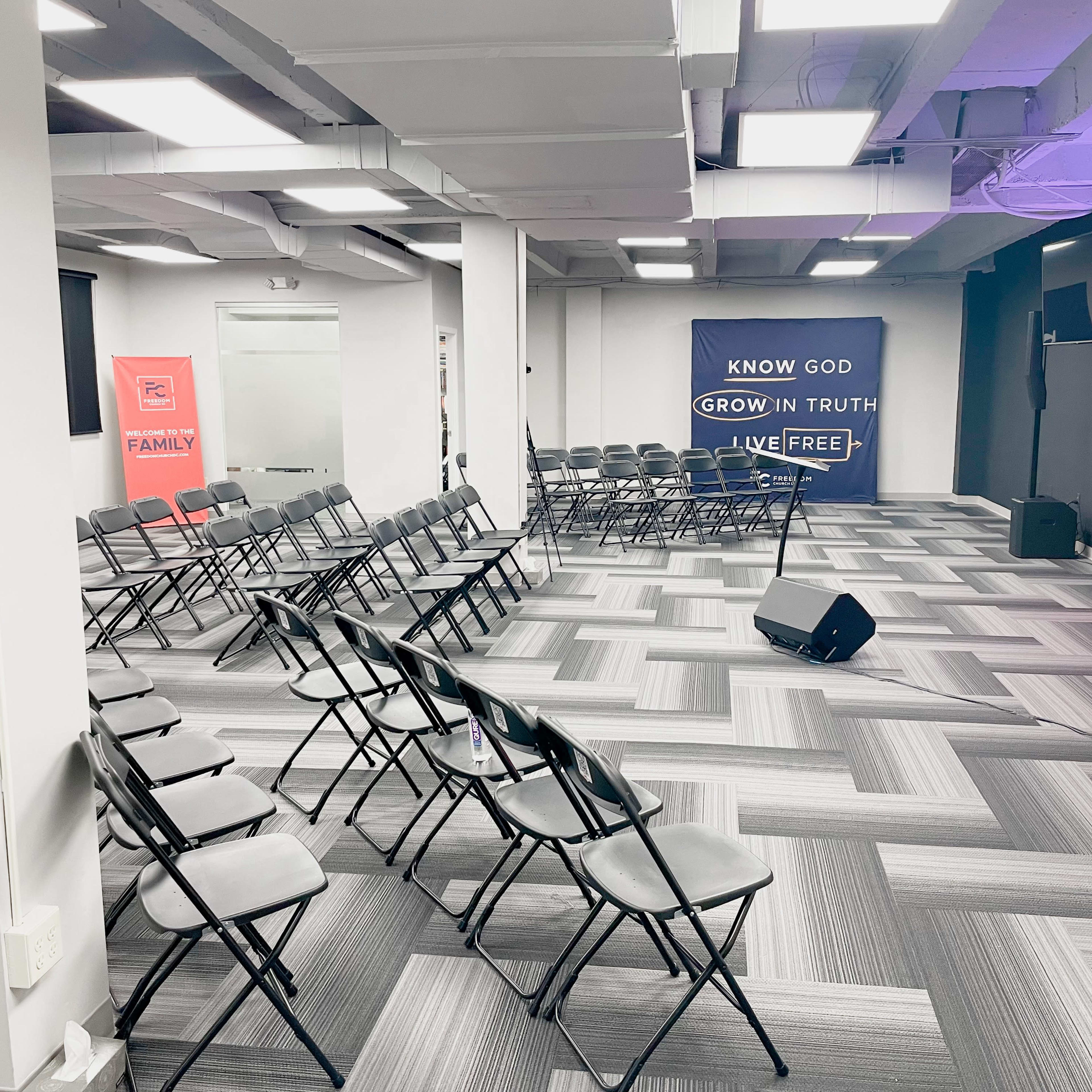 A spacious room arranged with rows of black folding chairs facing a stage with a microphone and banners on the walls.