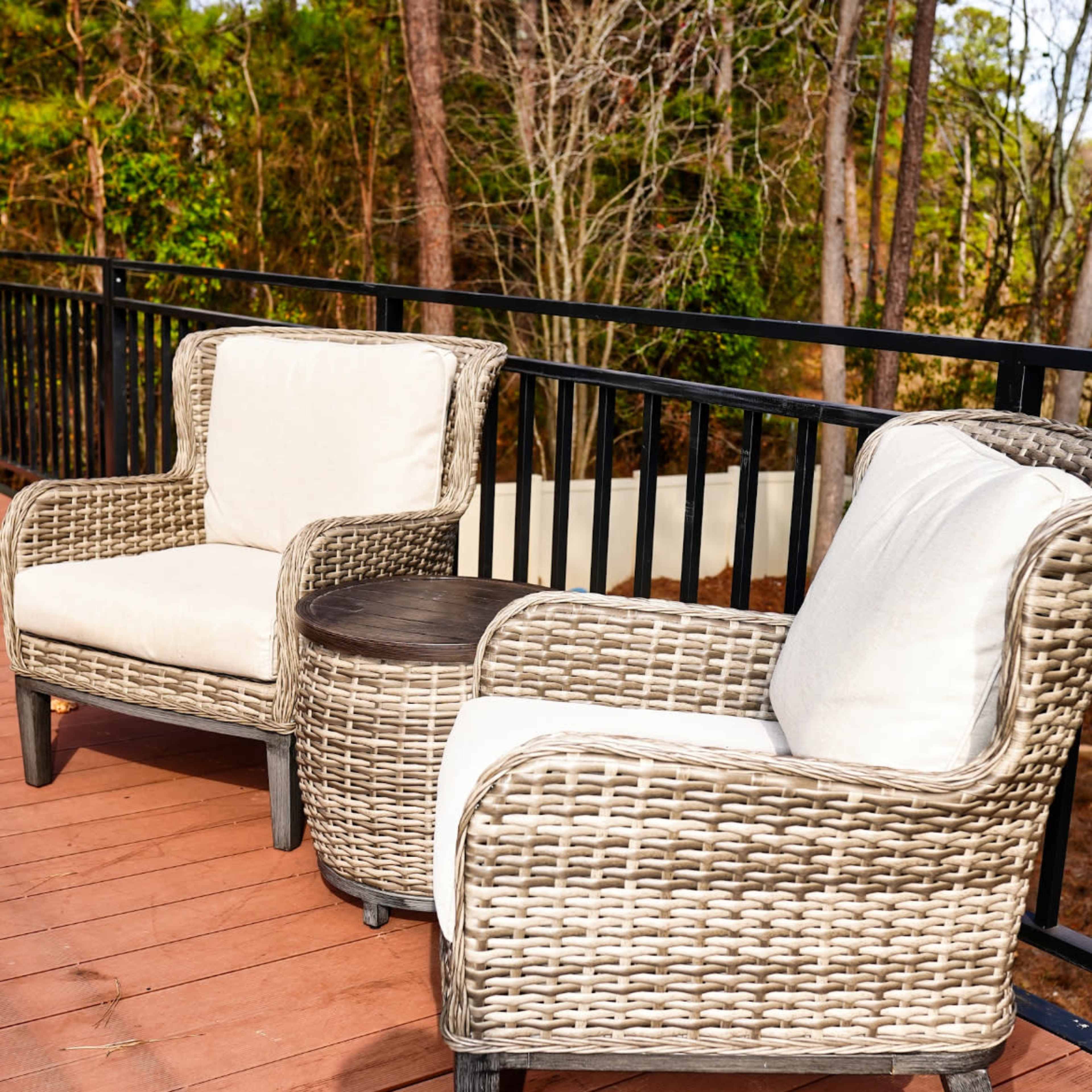Two wicker chairs with white cushions and a small round table are positioned on a wooden deck surrounded by trees.