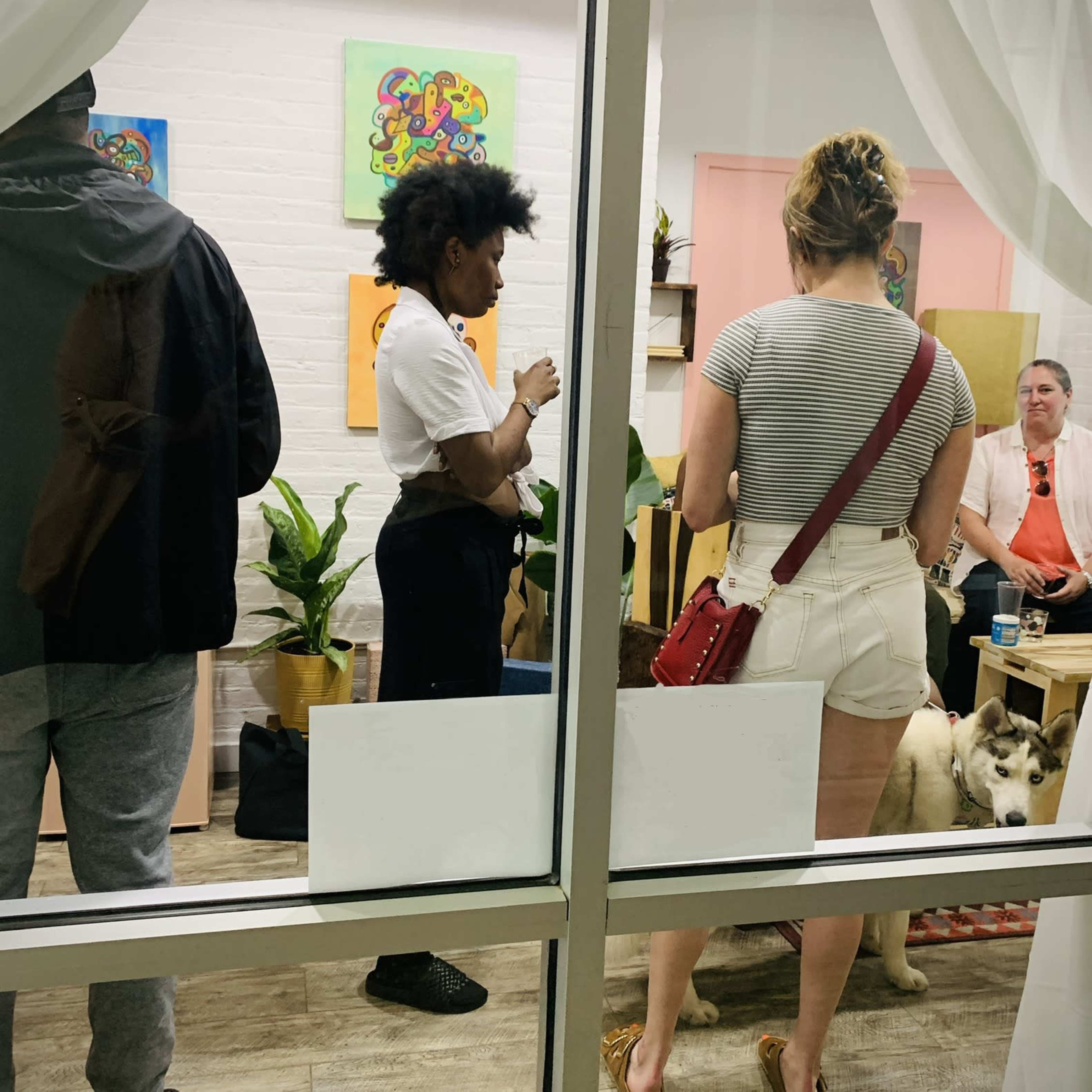 A gathering of people inside a room with plants, paintings on the walls, and a dog lying on the floor can be seen through a glass window.
