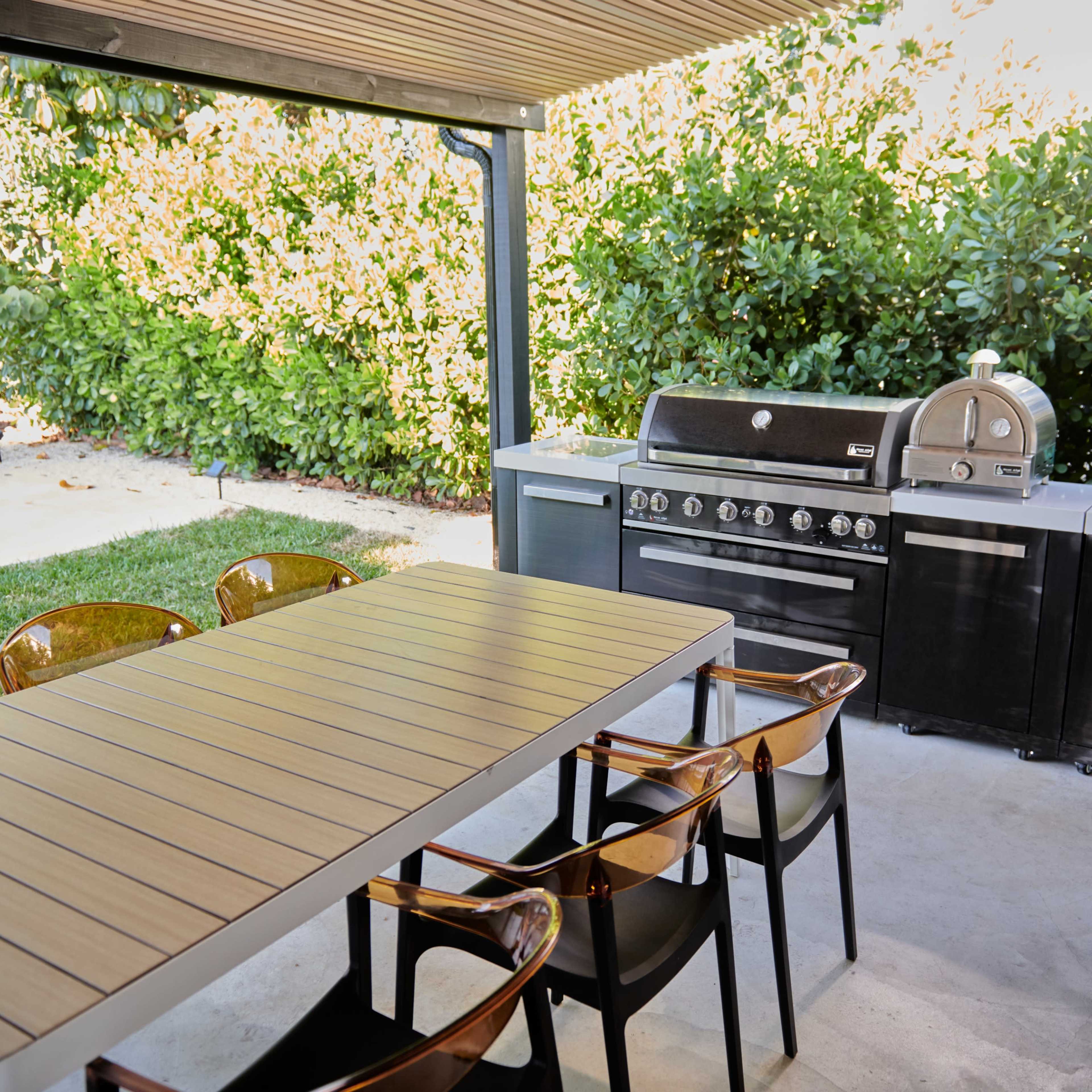 An outdoor kitchen area featuring a grill, a pizza oven, a sink, and a dining table with chairs under a covered structure, surrounded by greenery.