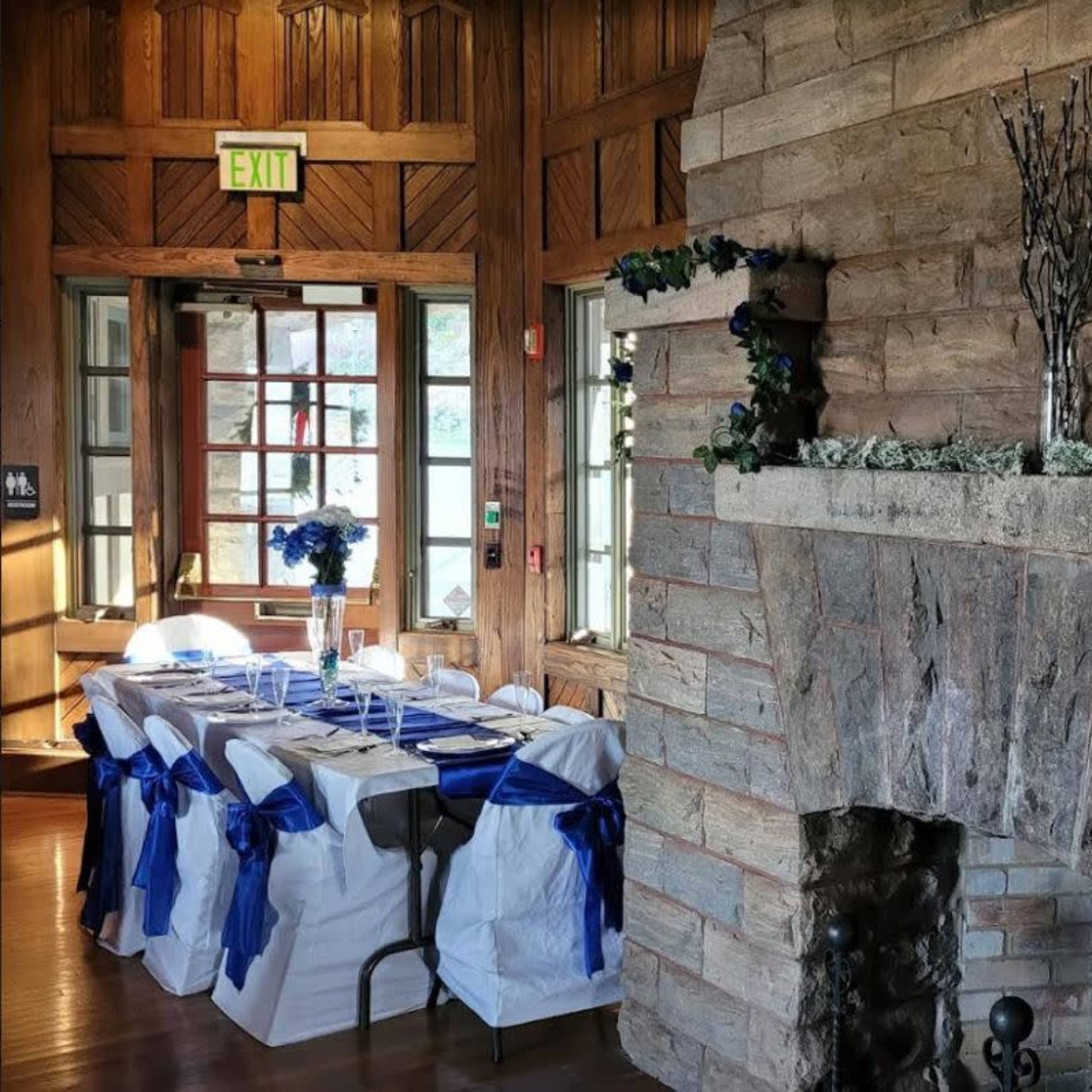 A dining area with a long table set for a meal, adorned with blue decorations, next to a stone fireplace.
