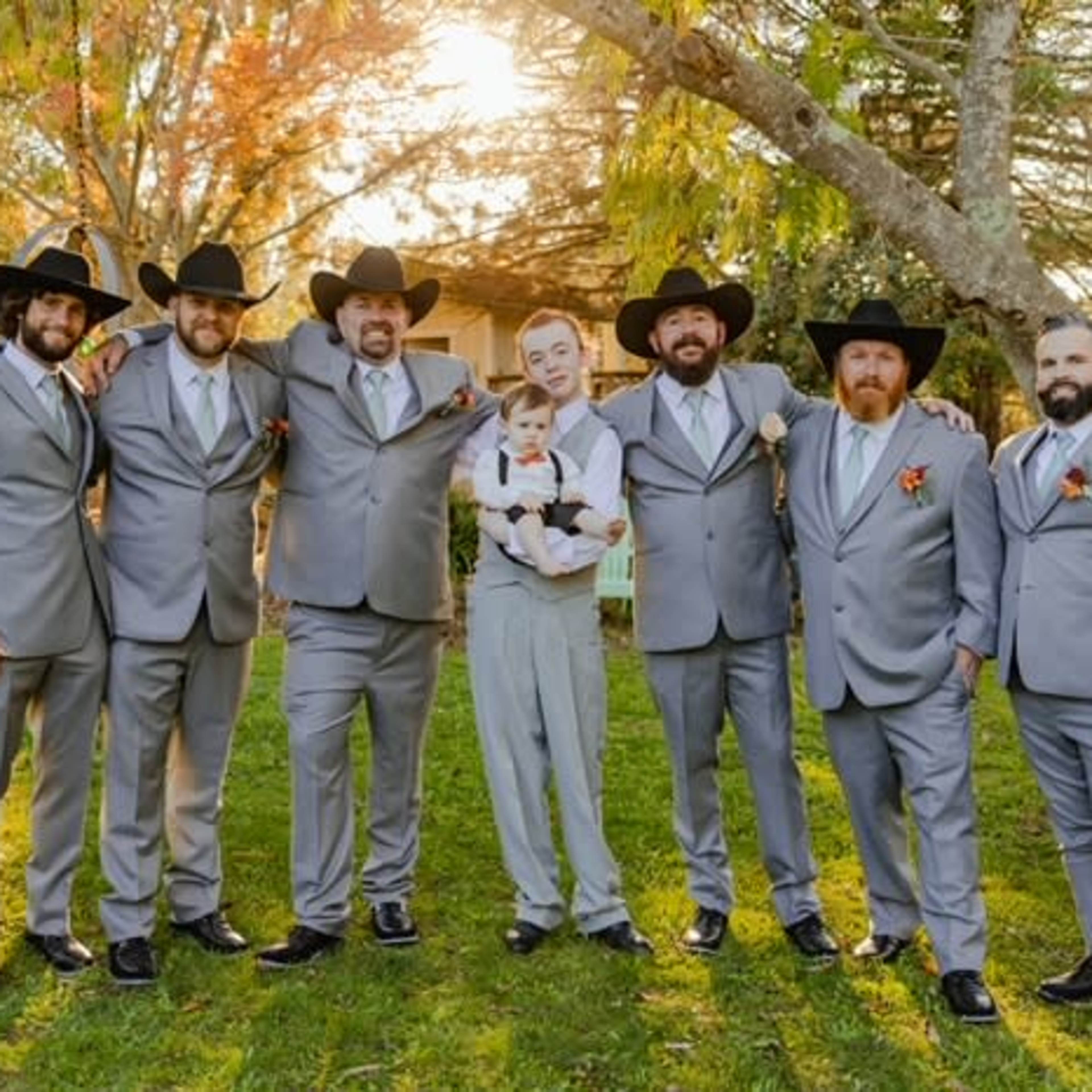 A group of seven men wearing gray suits and black hats stands together outdoors, with one man holding a baby, as sunlight filters through the trees behind them.