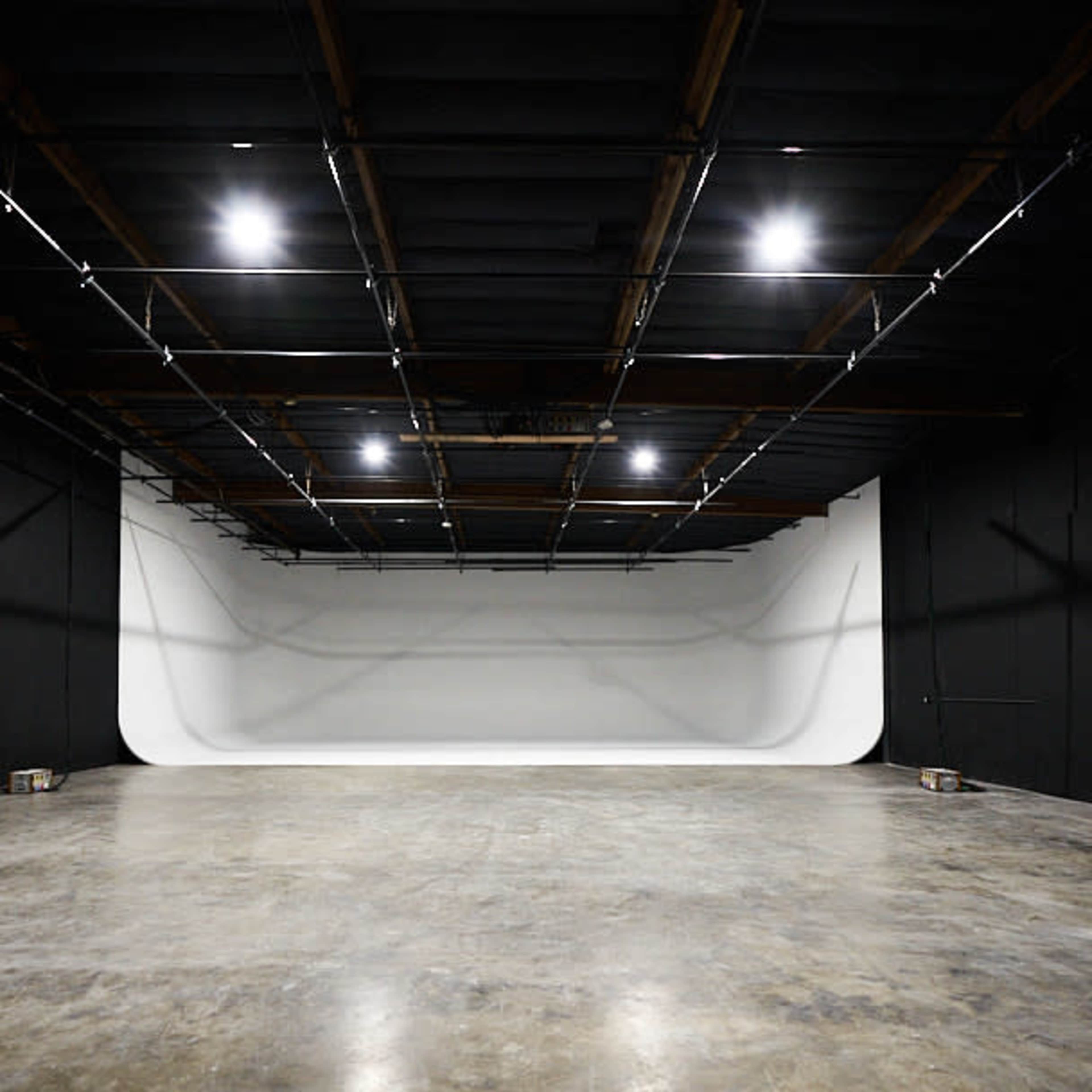 A spacious studio with a seamless white backdrop at one end and dark walls surrounding a concrete floor.