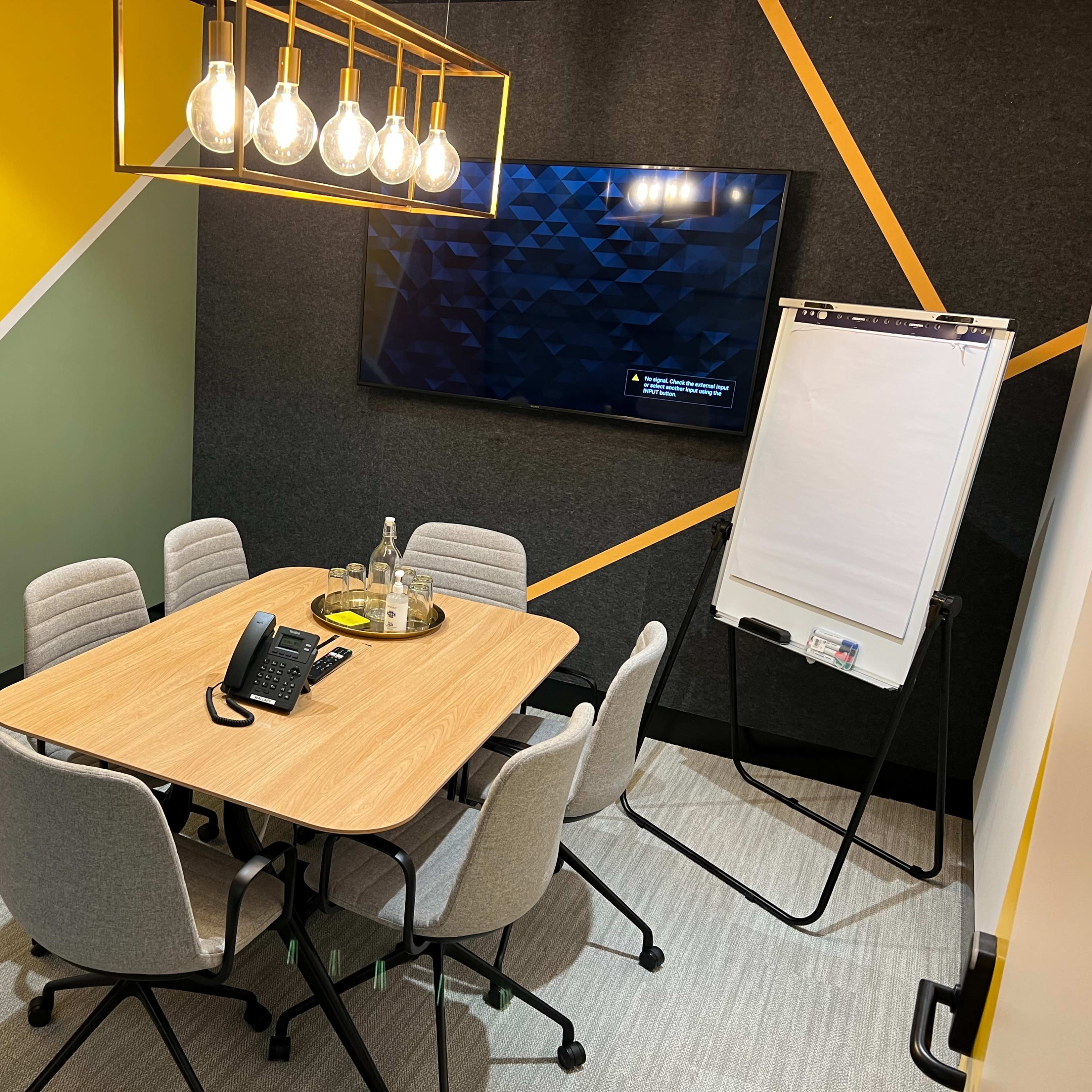 A small meeting room with a wooden table, six chairs, a large screen on the wall, and a whiteboard on a stand.