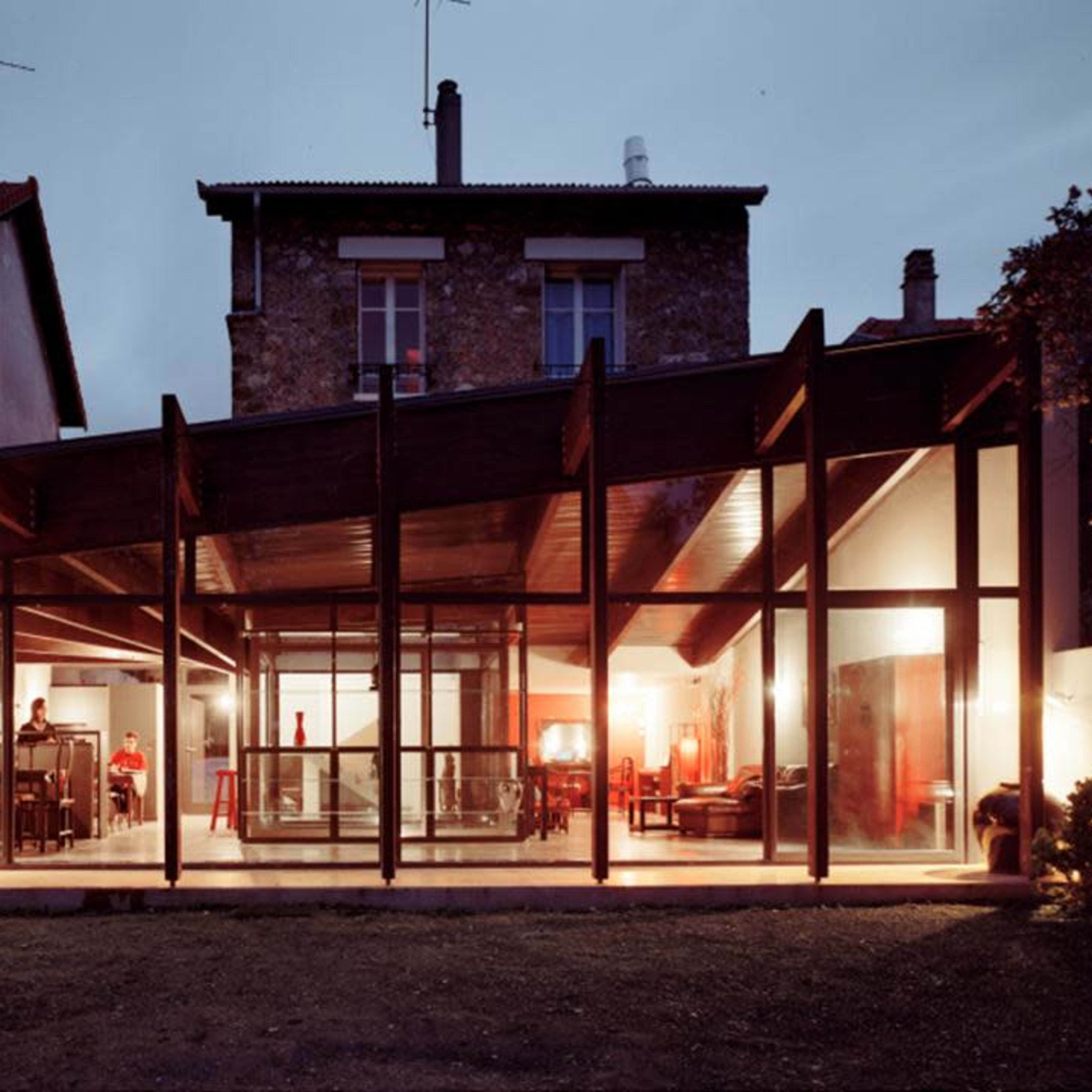 A modern house with a glass facade and wooden structure is illuminated at dusk, showcasing an open interior space.
