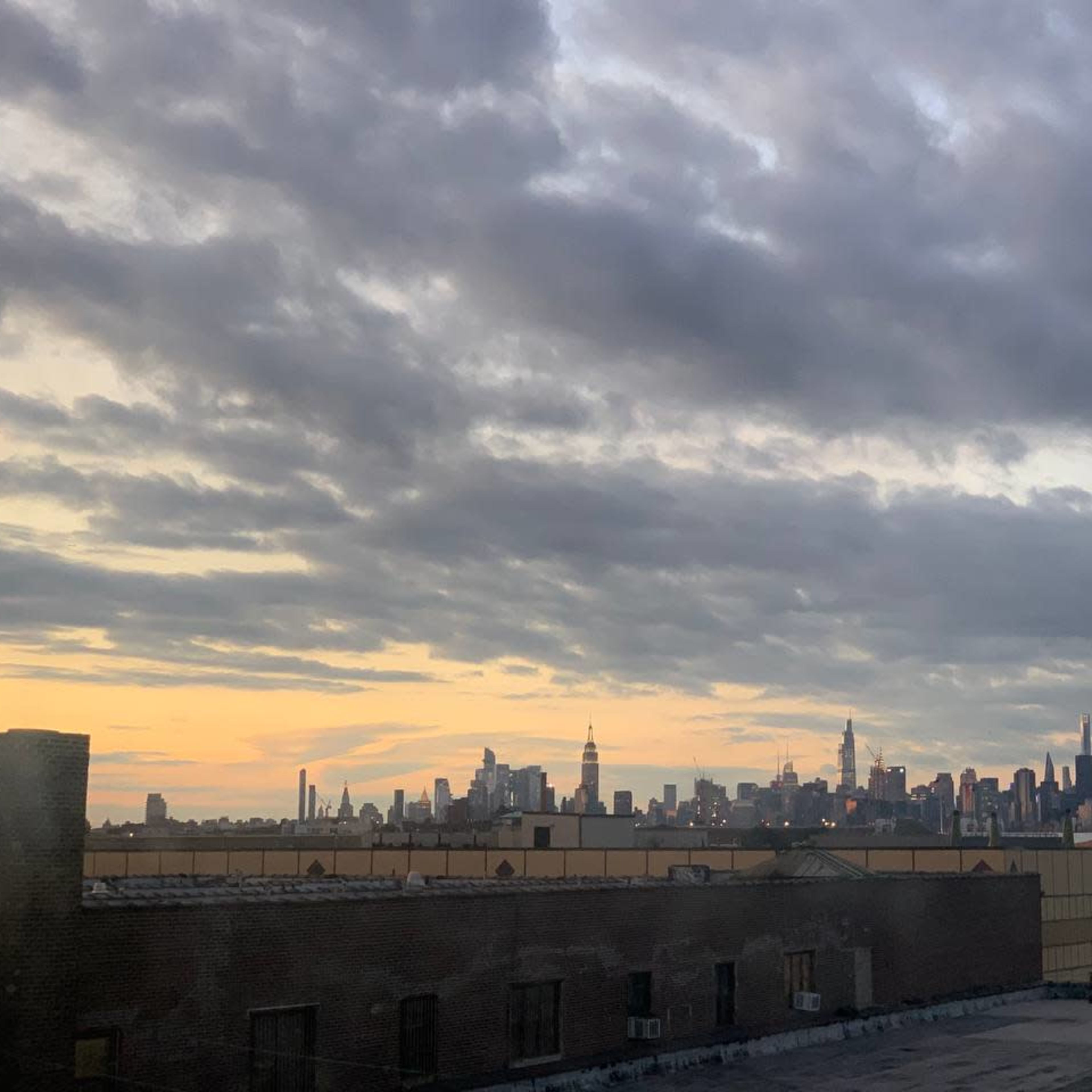 The skyline of a city is visible against a backdrop of clouds and a fading sunset, with various tall buildings, including the Empire State Building, rising in the distance.