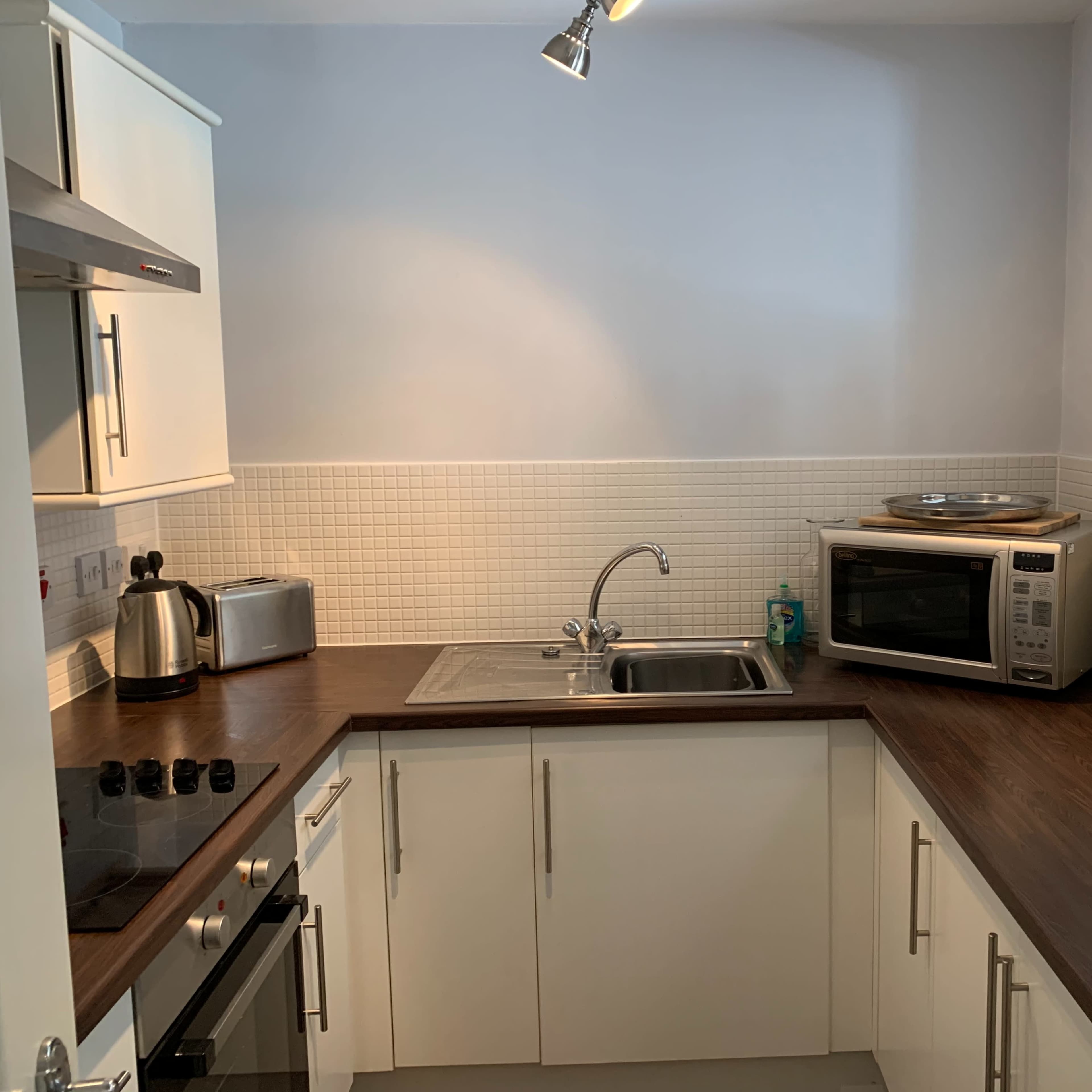 A kitchen with white cabinetry, a stainless steel sink, a microwave, and a countertop with a stove and kettle.