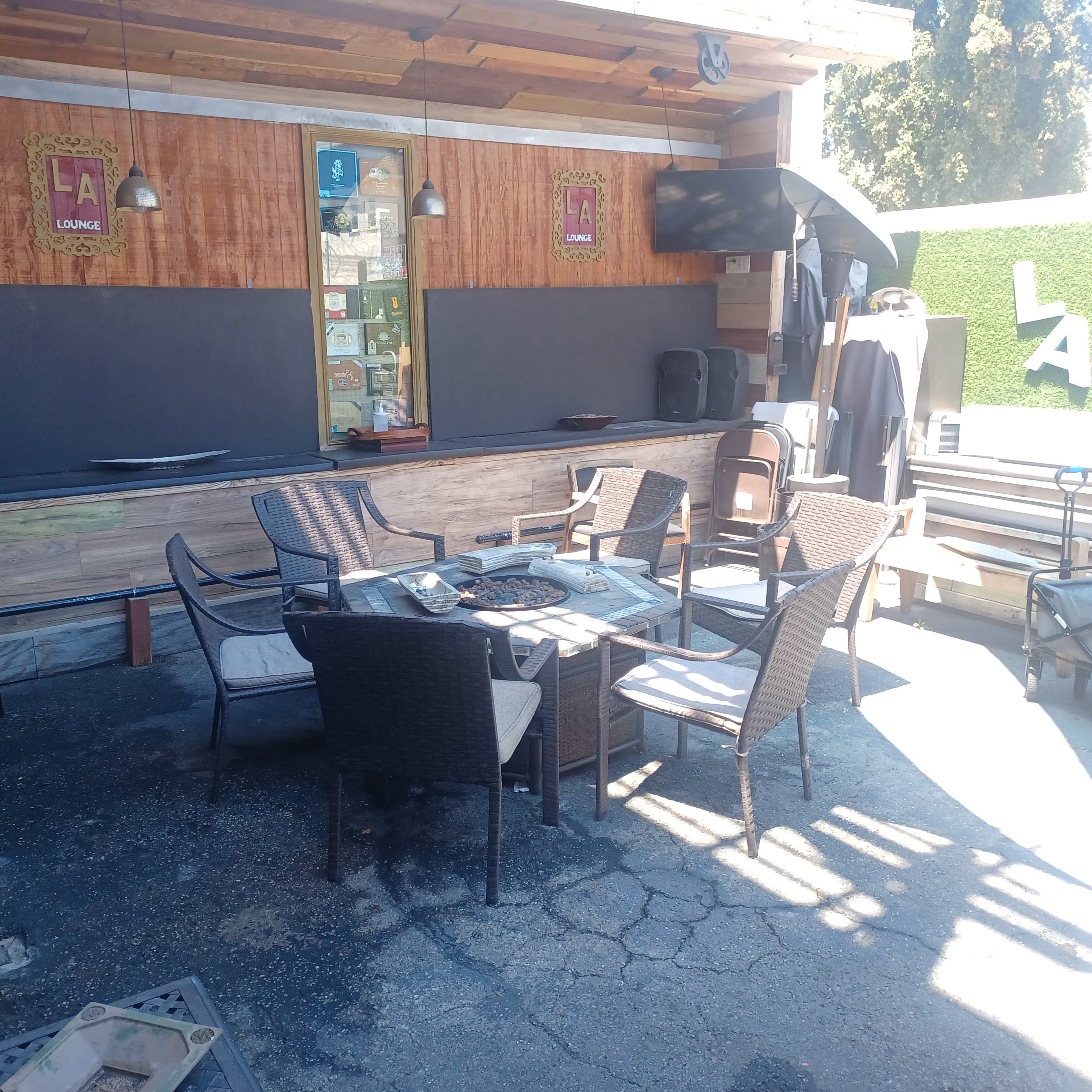 An outdoor lounge area with a table and several chairs arranged around it, adjacent to a bar counter and a backdrop featuring green plants.