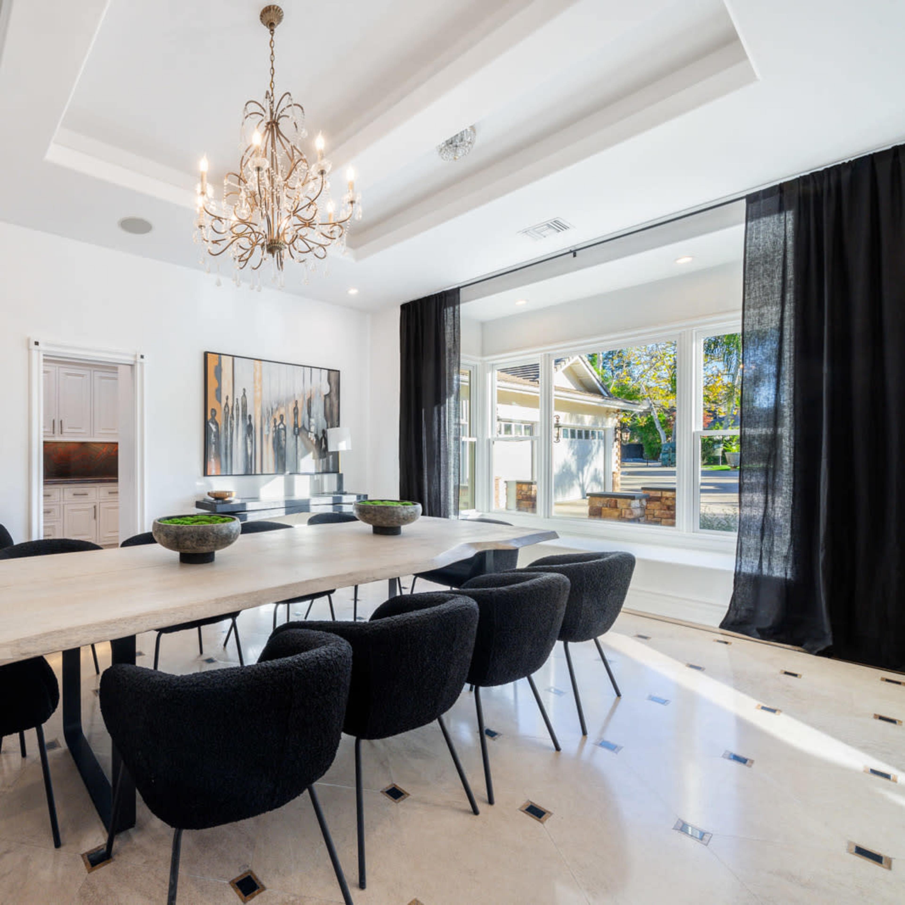 A large dining table surrounded by black upholstered chairs is set in a bright room with a chandelier and a window overlooking an outdoor space.