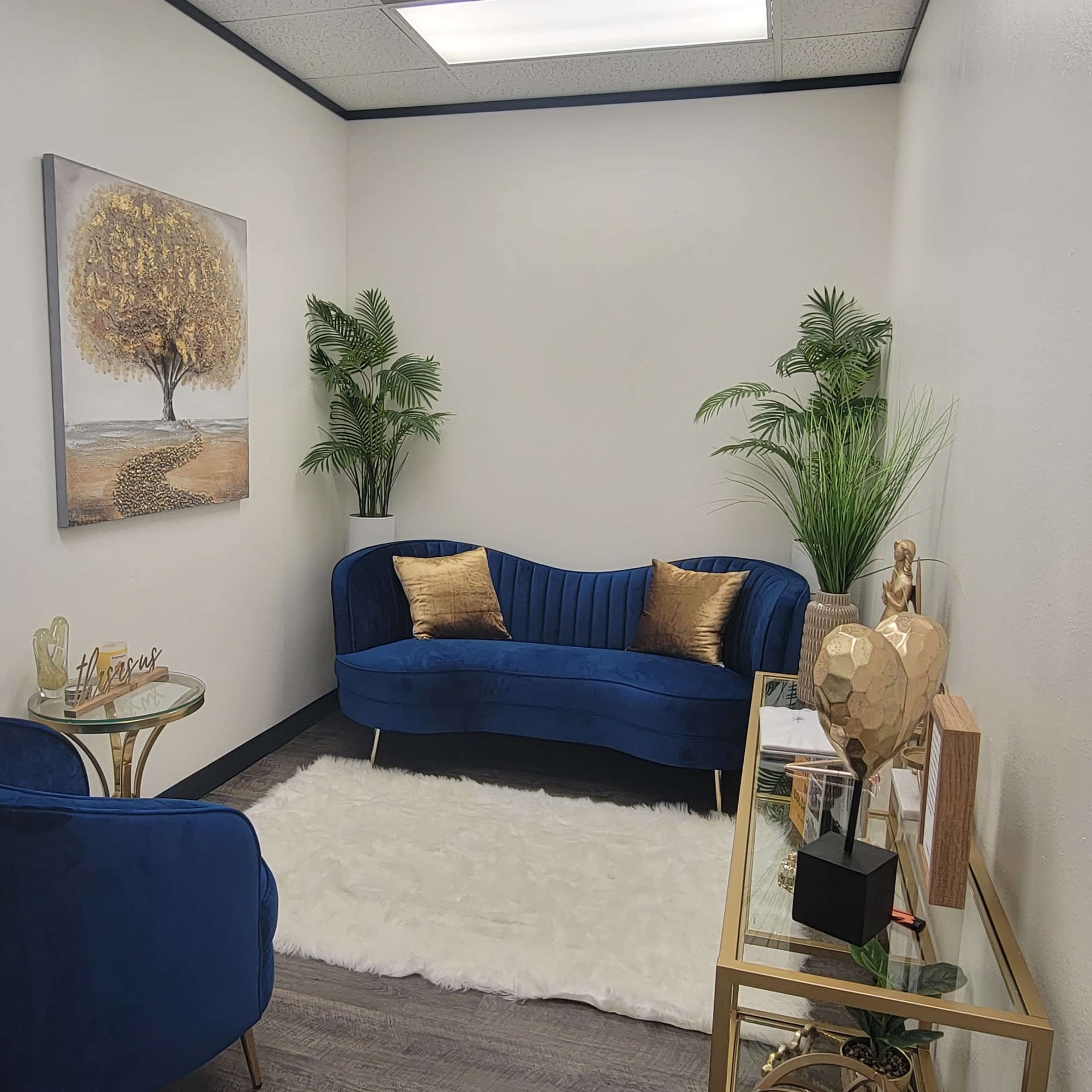 A small, elegantly arranged waiting area with a blue upholstered sofa, a matching chair, decorative plants, and a wall art featuring a tree.