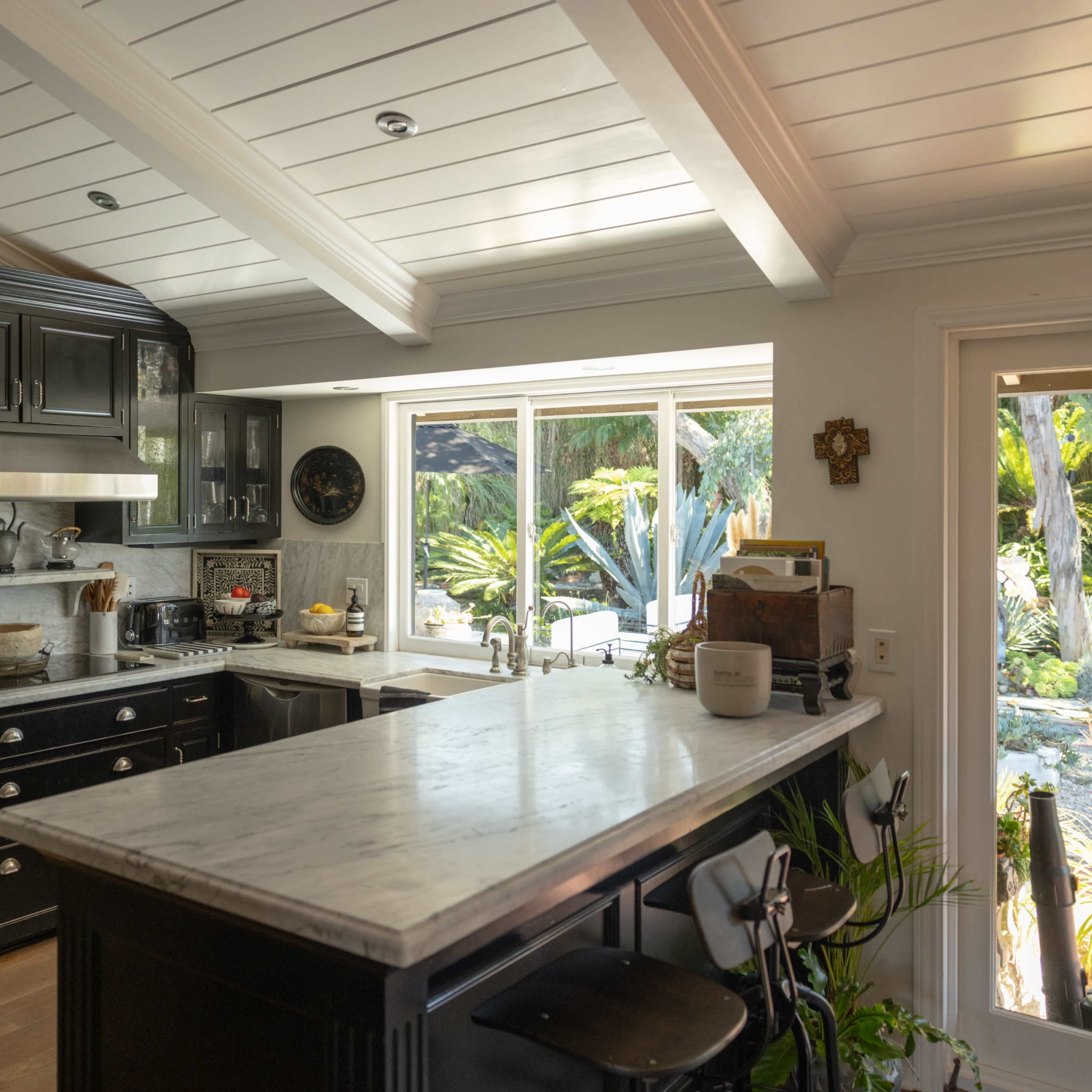 A modern kitchen features black cabinetry, a marble countertop, and a large window with views of a garden.