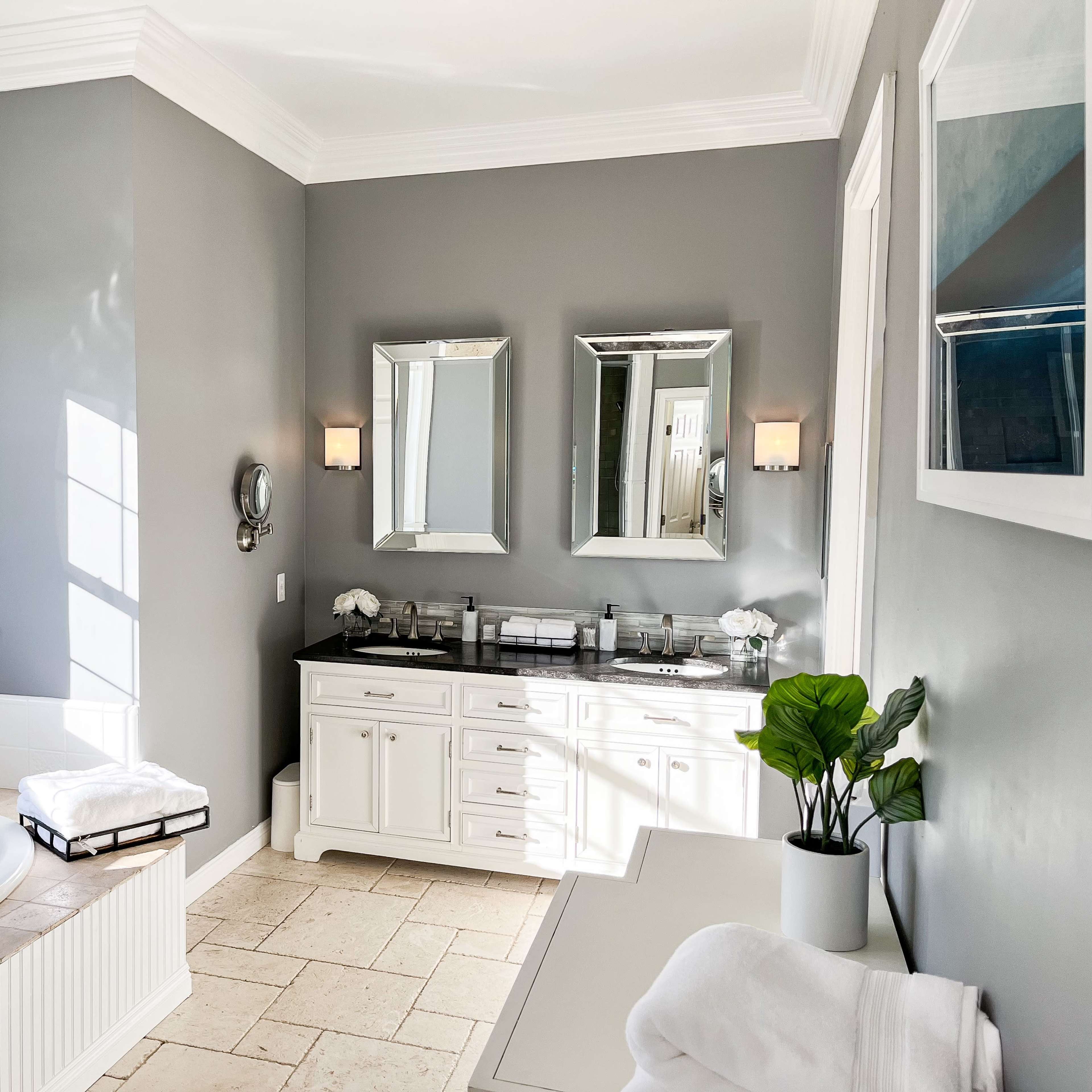 The bathroom features a large jetted tub beside a vanity with double mirrors and sinks, all set against gray walls and stone flooring.