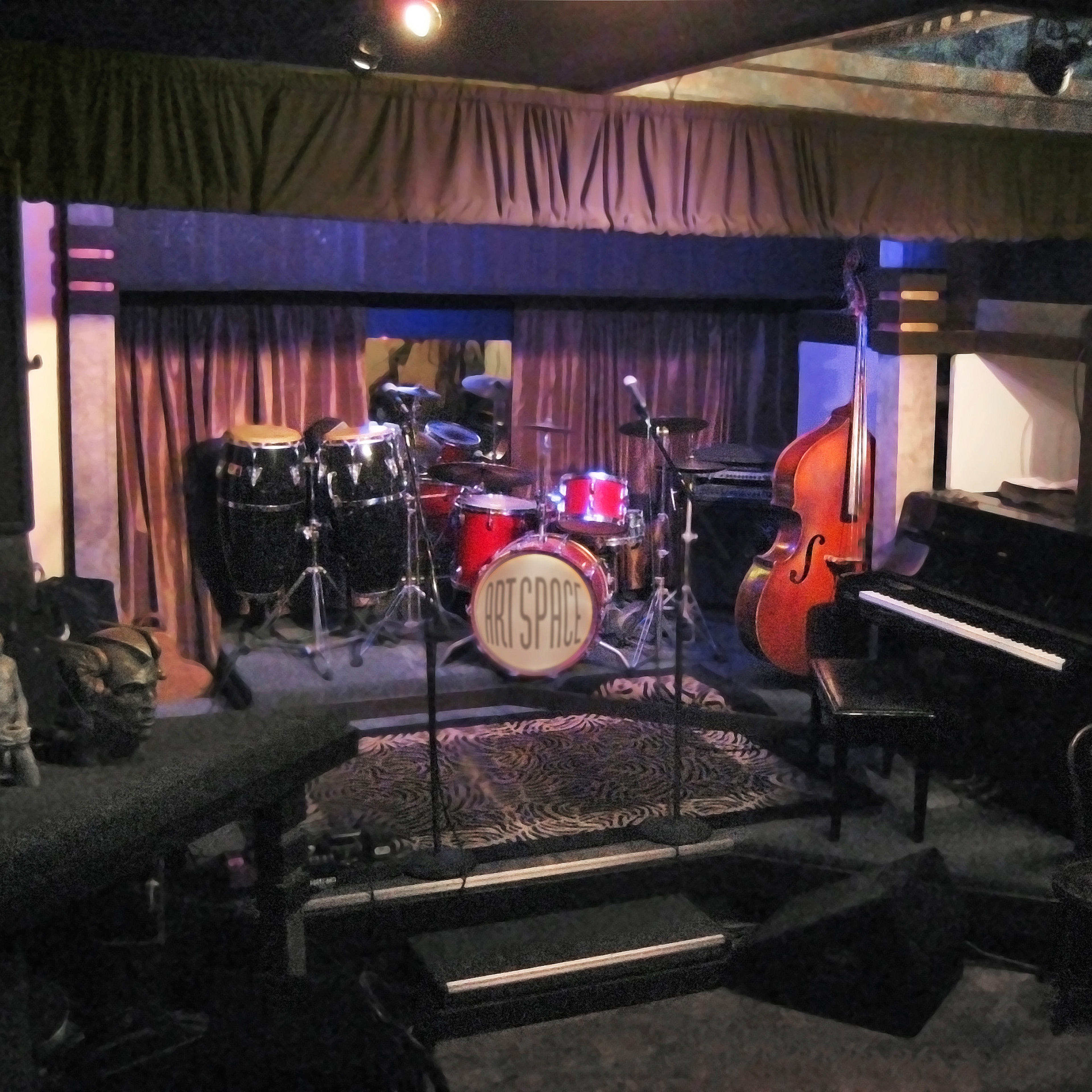 A stage set up for a performance, featuring a drum kit, a double bass, and a piano, with decorative elements in a dimly lit venue.