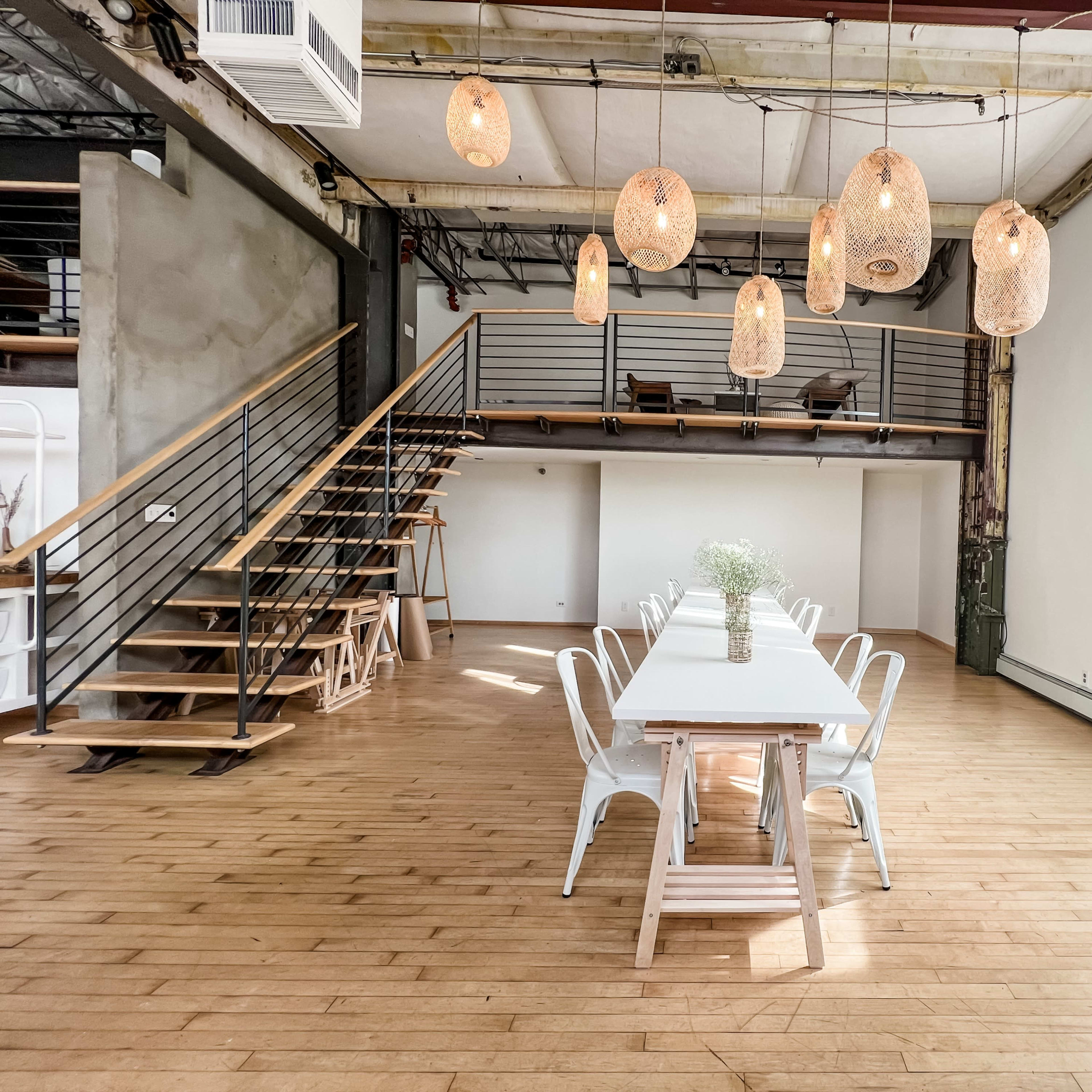 A spacious room features a long dining table with a vase at its center, surrounded by white chairs, and is illuminated by hanging pendant lights, with a staircase leading to a second level in the background.