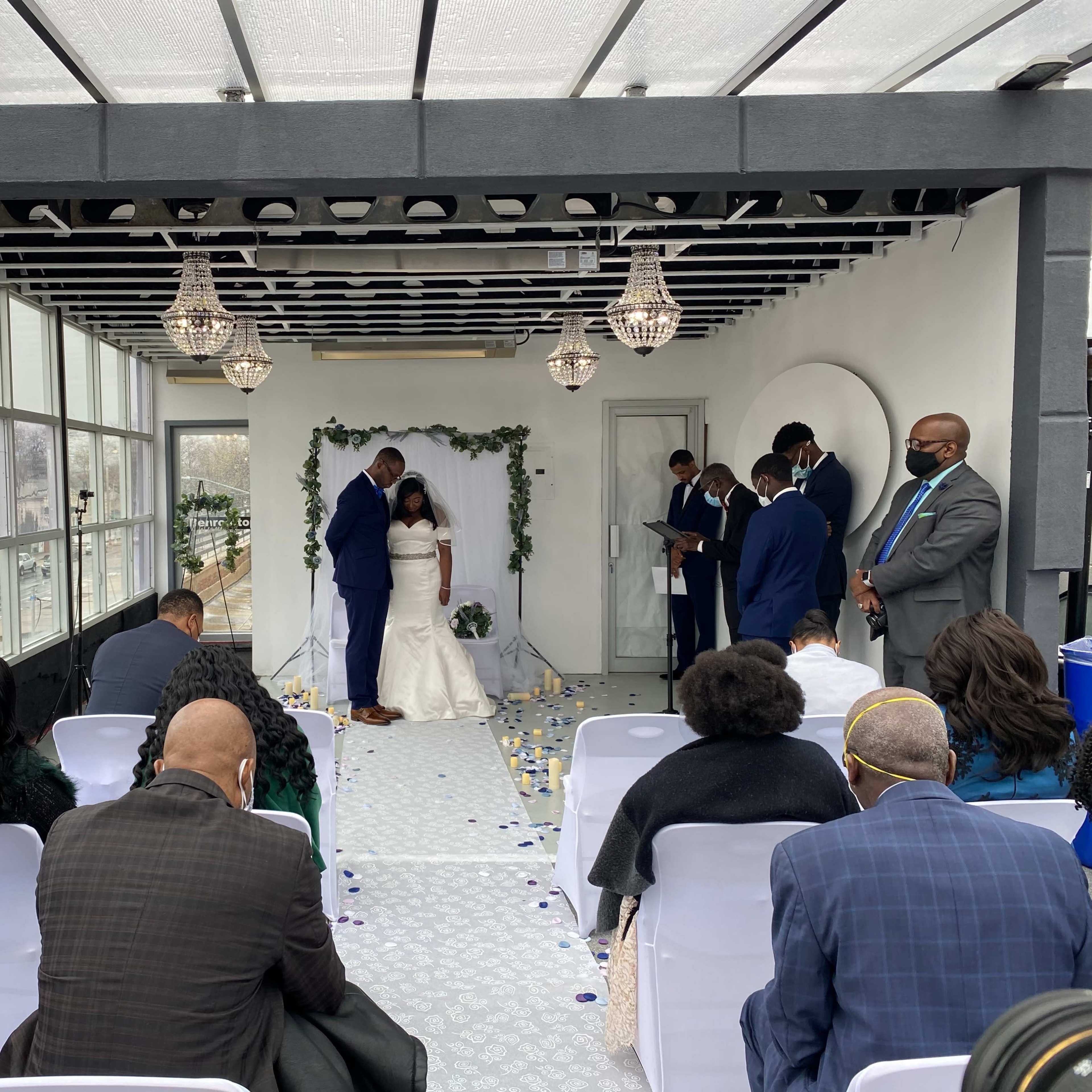 A couple stands at the altar during a wedding ceremony, surrounded by guests seated in white chairs, with floral decorations and overhead lights in a glass-enclosed space.
