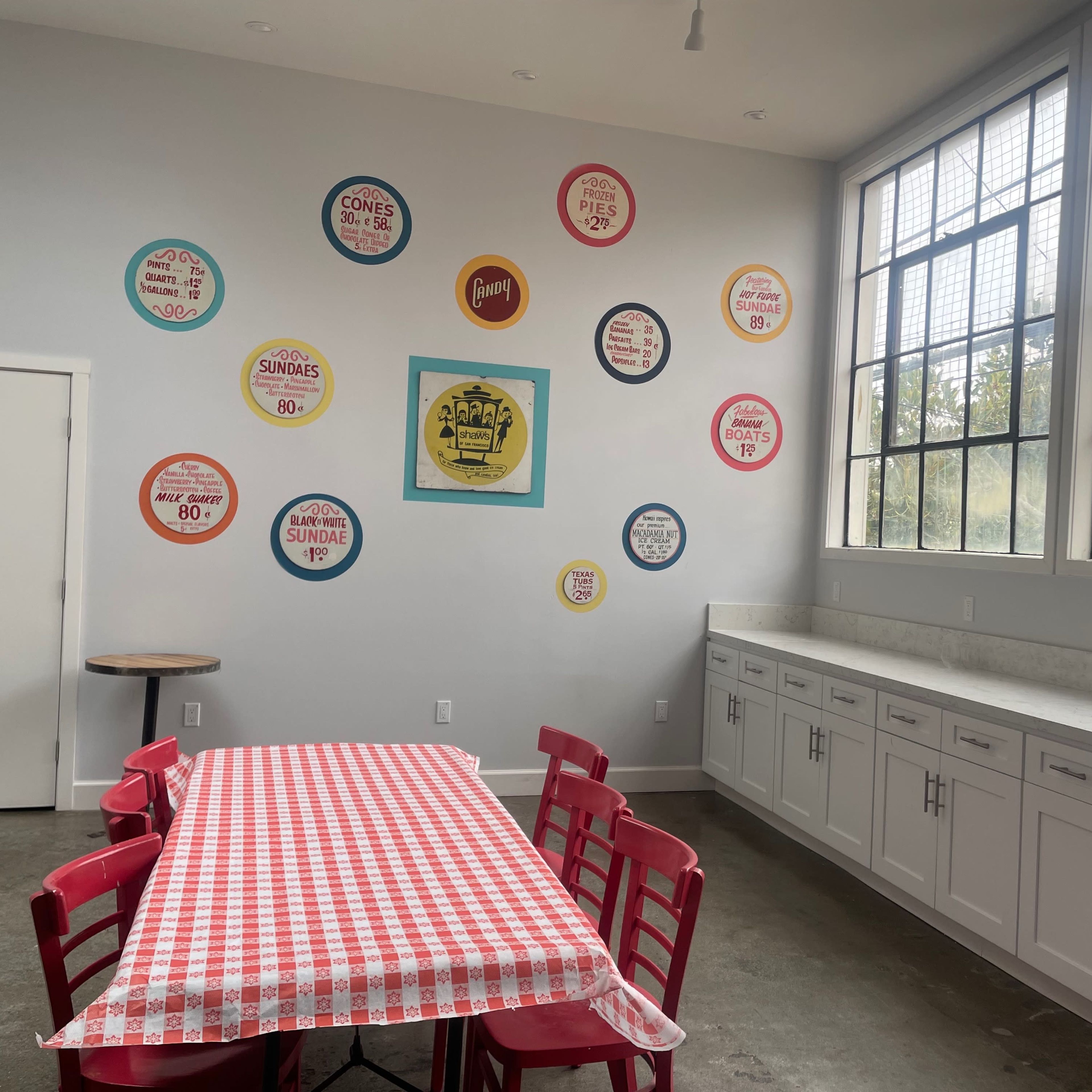 A bright room features a red-checkered table surrounded by red chairs, with vintage-style signs displayed on the wall.