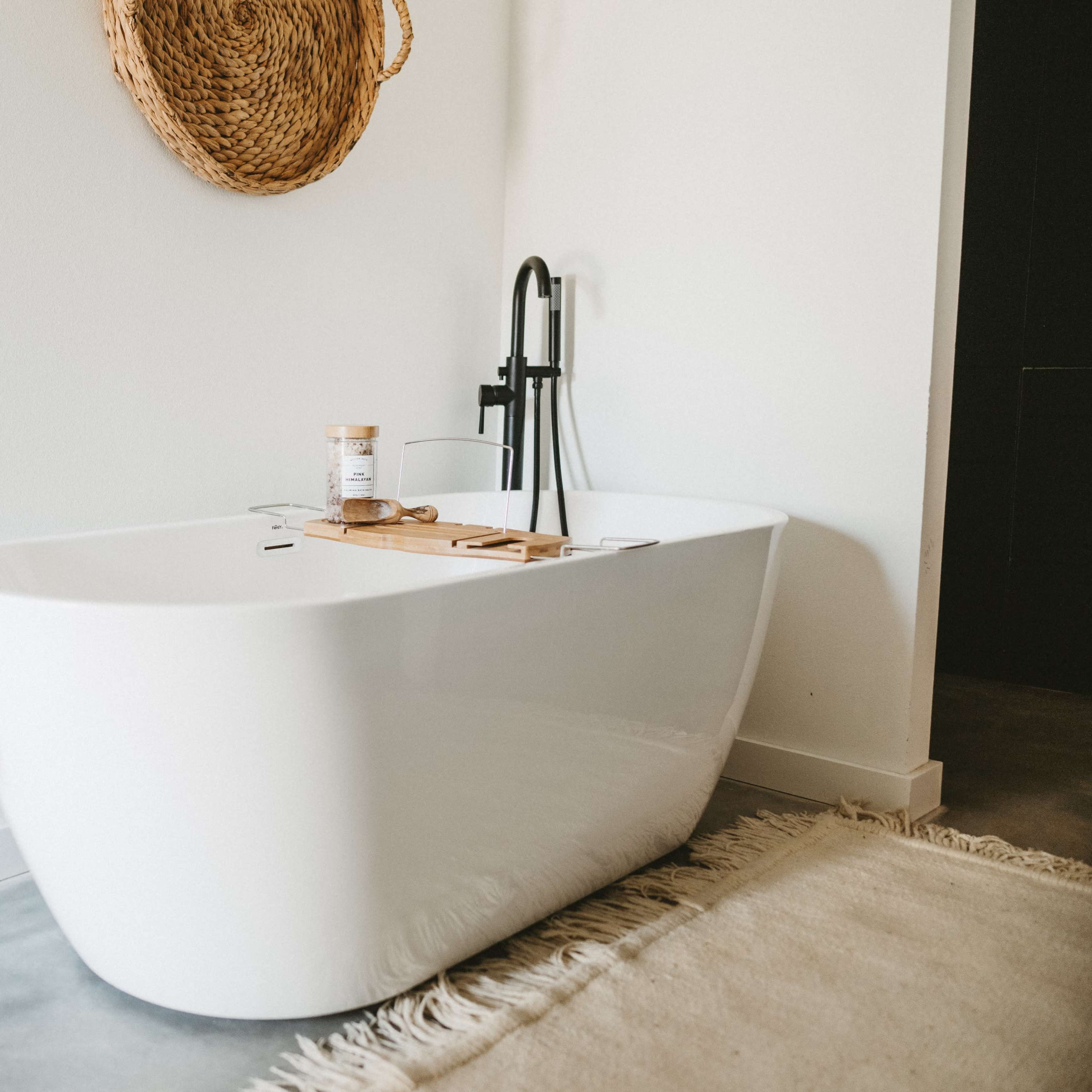 A modern freestanding bathtub beside a wooden side table with a glass of water and a natural fiber rug on the floor.